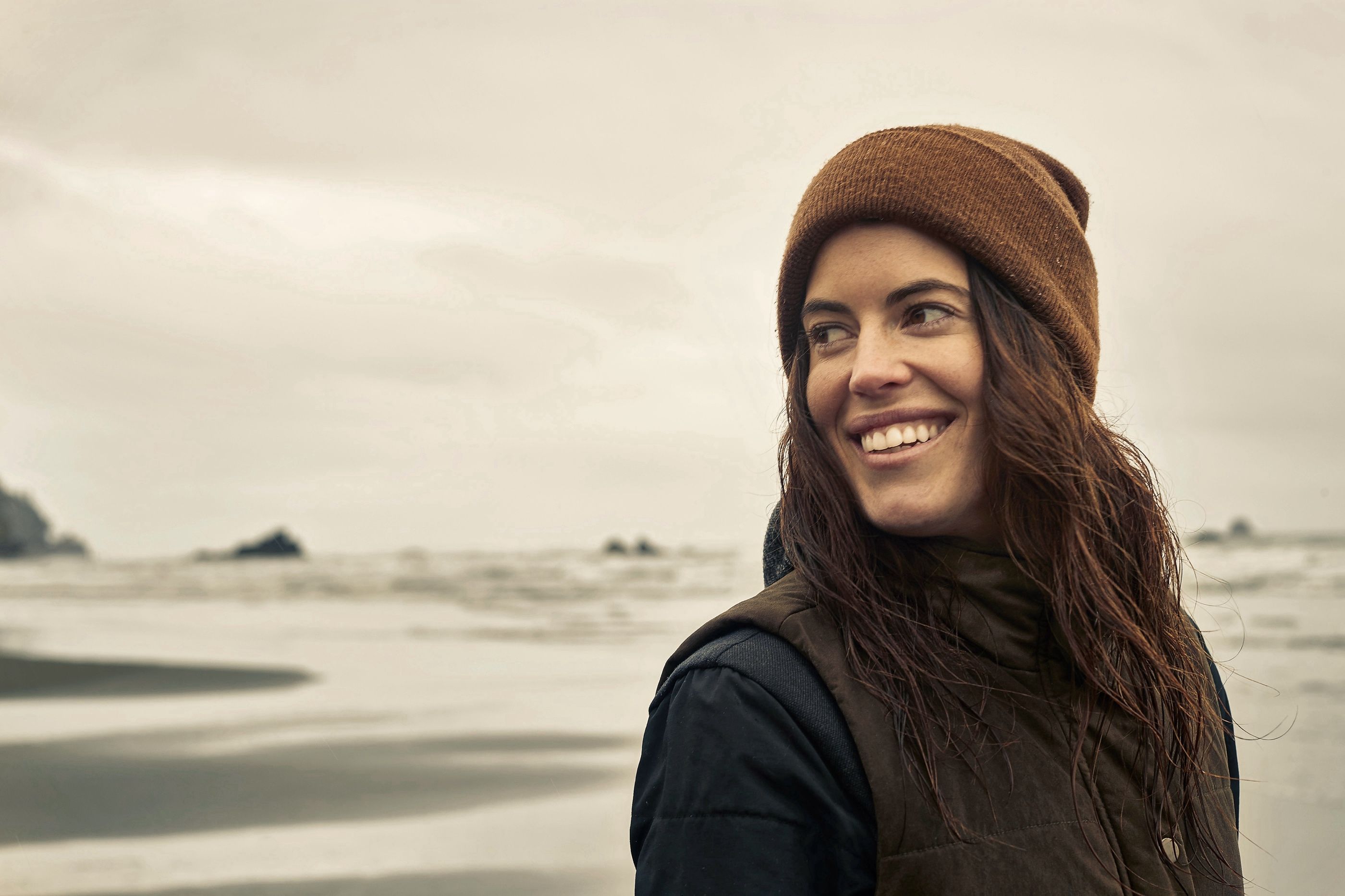 Woman in a brown beanie smiles on a beach with rock formations and an overcast sky in the background.