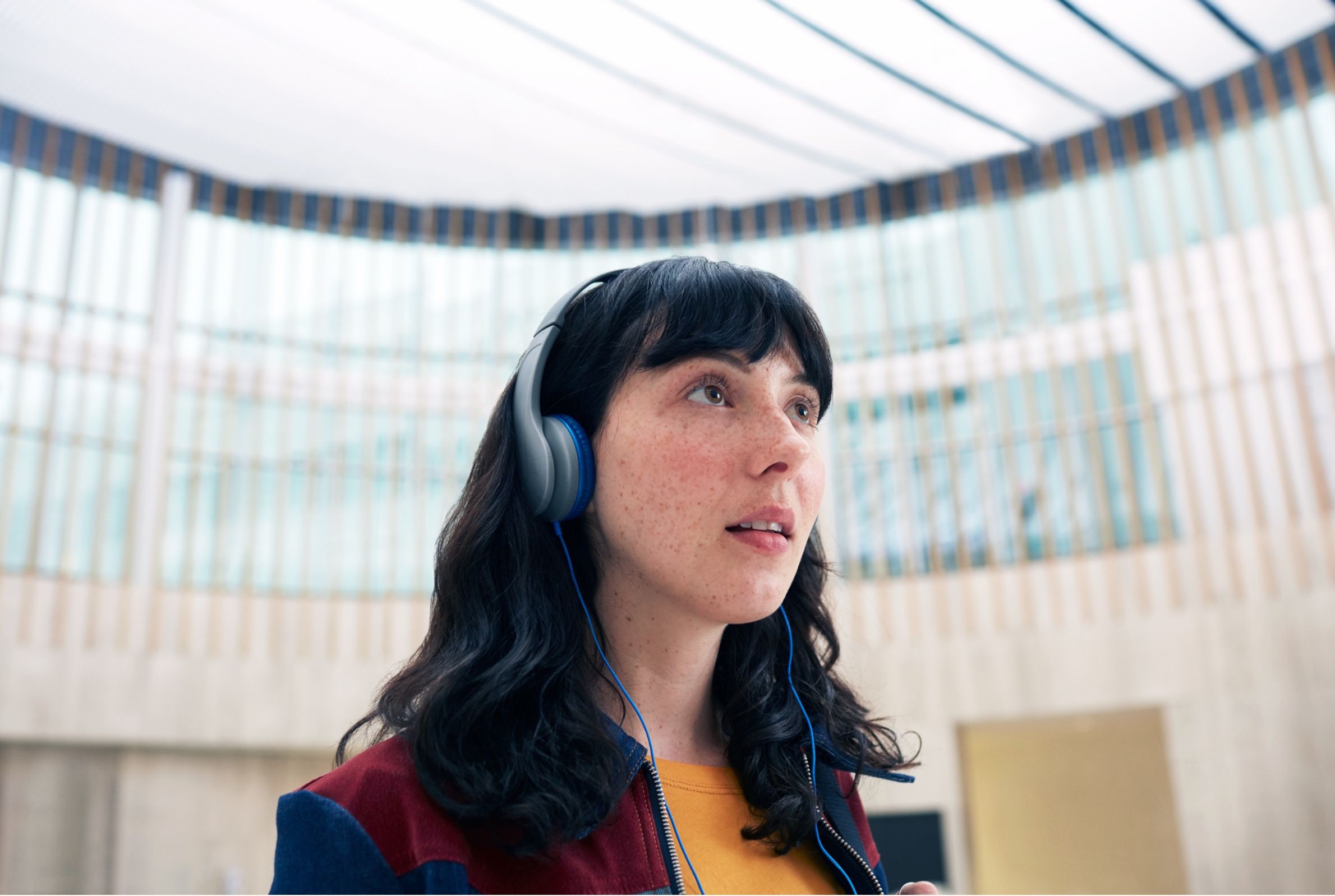 Woman wearing headphones looks upward inside a glass-walled atrium, wearing a colorful jacket.