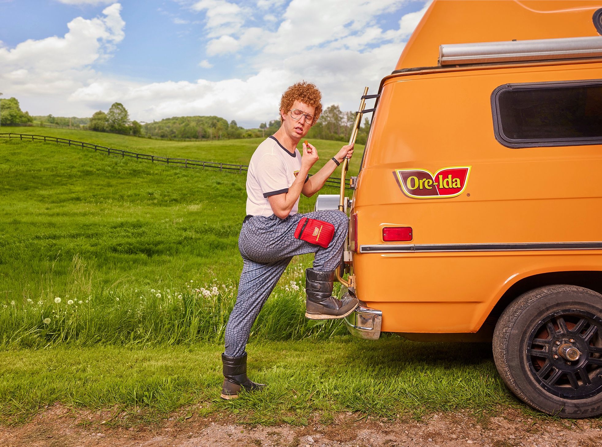 Man with curly red hair and glasses eats a fry, boot on an orange van's ladder; Ore-Ida logo visible on the van.