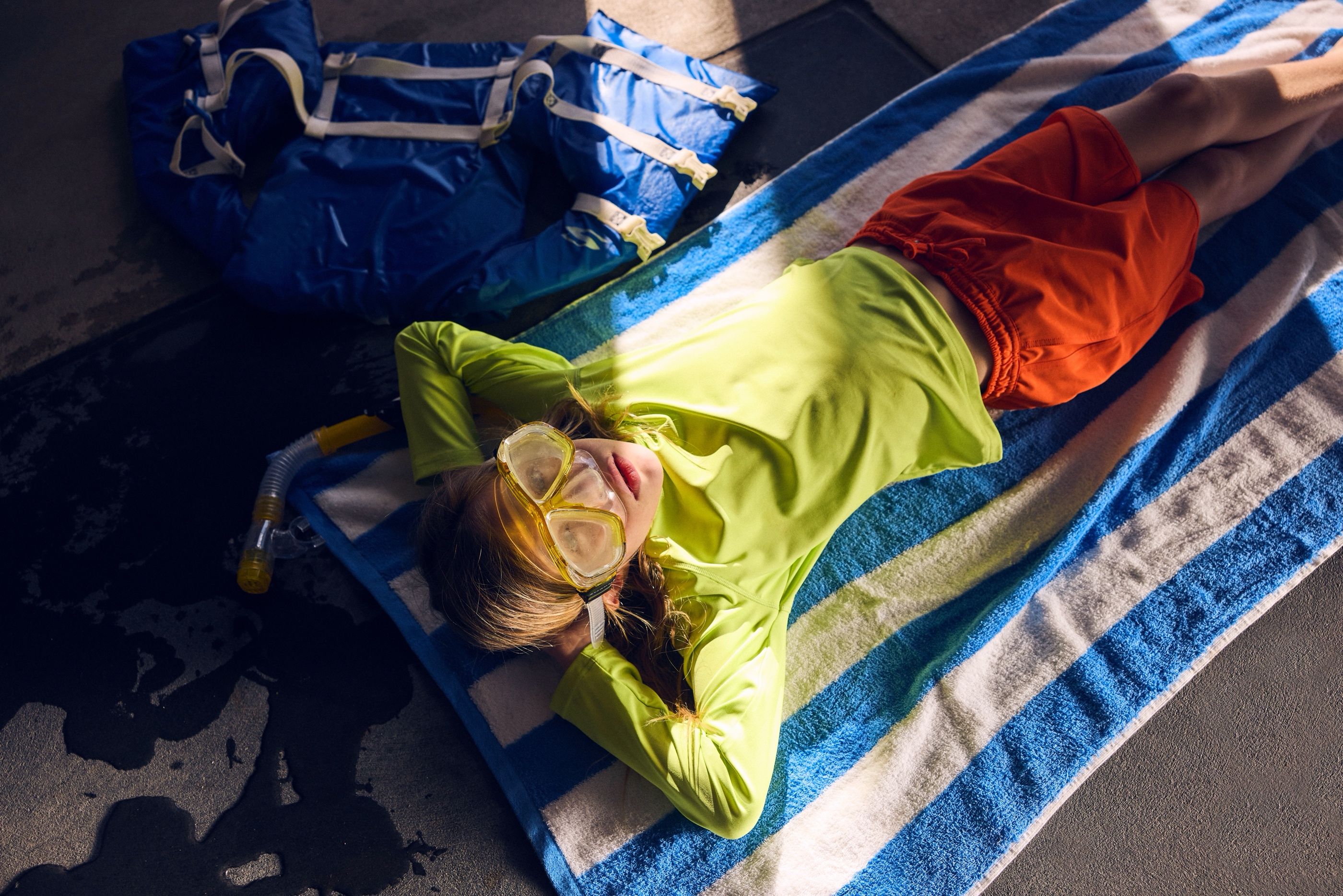 Child lying on a striped towel wearing a green rash guard and snorkel mask, with a life vest bag nearby.