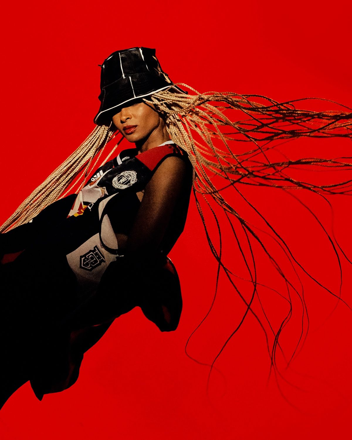 Woman in profile wearing a black bucket hat and Manchester United jersey, long blonde braids, red backdrop.