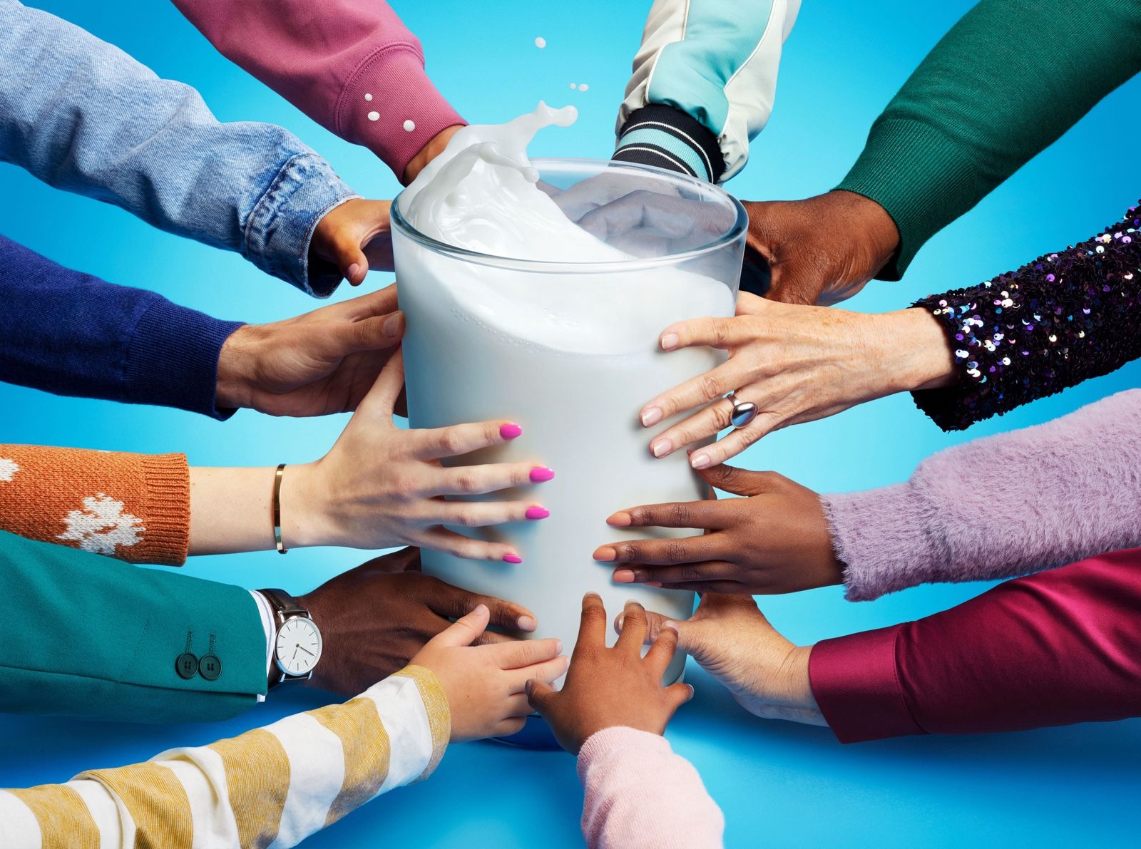 Multiple hands in colorful sleeves reach into a large glass of milk splashing over the rim, on a blue background.