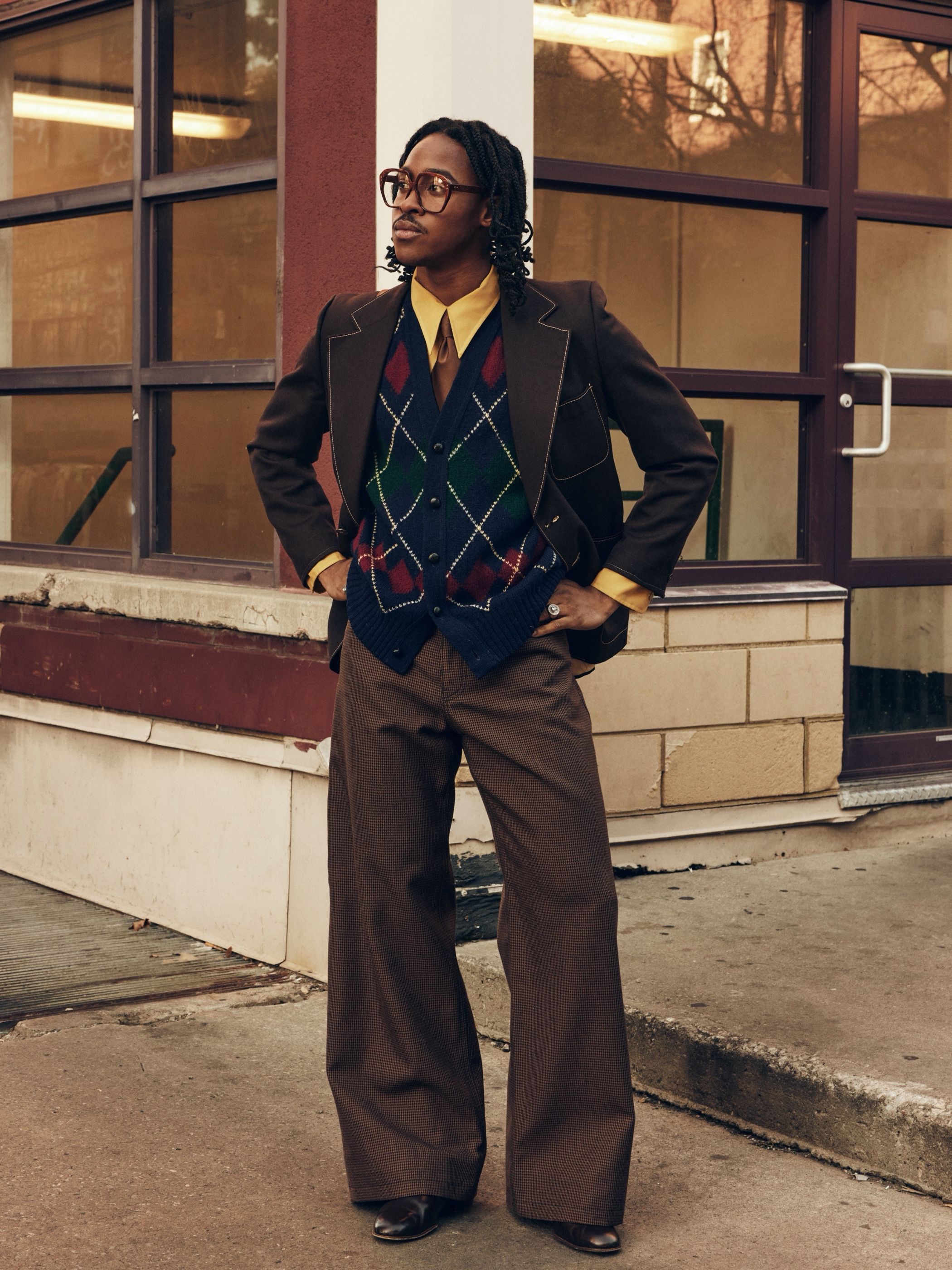Man with braided hair wears a brown blazer over a navy argyle cardigan, mustard shirt and tie, and wide-leg trousers.