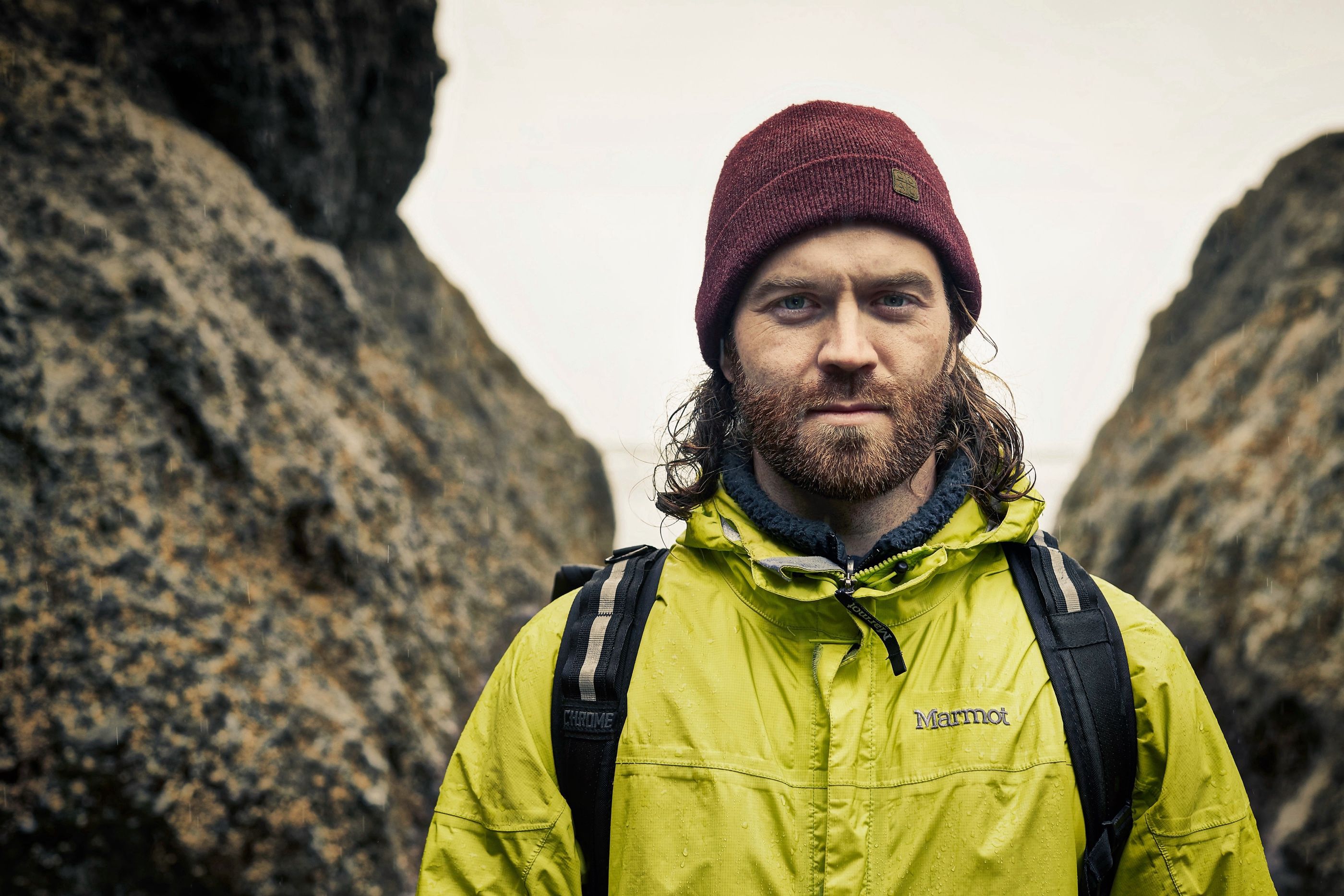 Bearded man in a maroon beanie and yellow Marmot rain jacket stands between large rocks, backpack visible.
