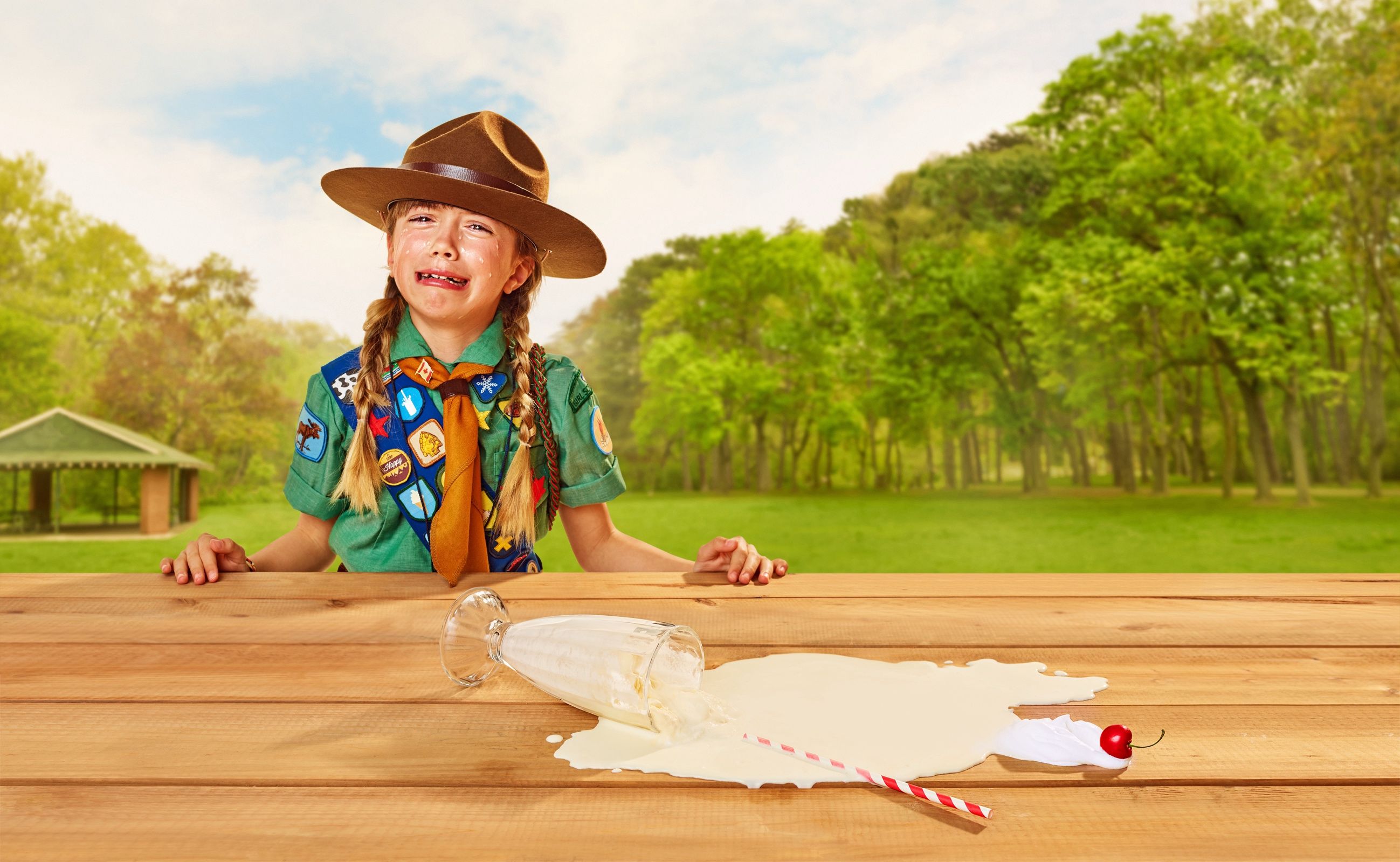 Girl in a scout uniform and campaign hat cries behind a picnic table with a spilled milkshake in a park.