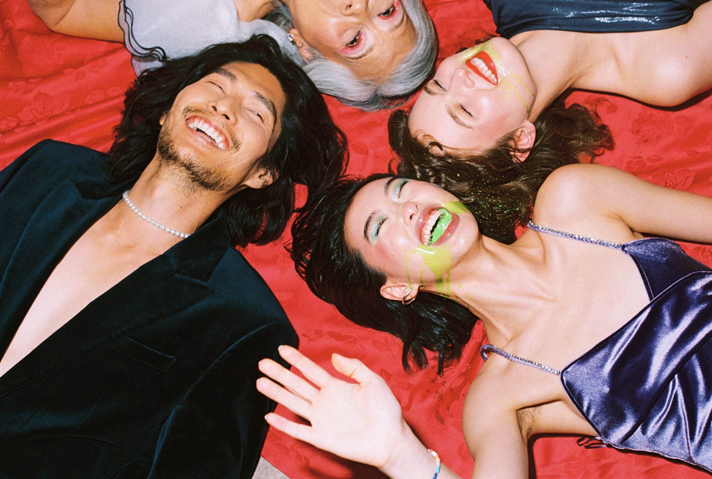 Four people lying face-up in a circle, laughing, with green paint or icing dripping on one person's face.