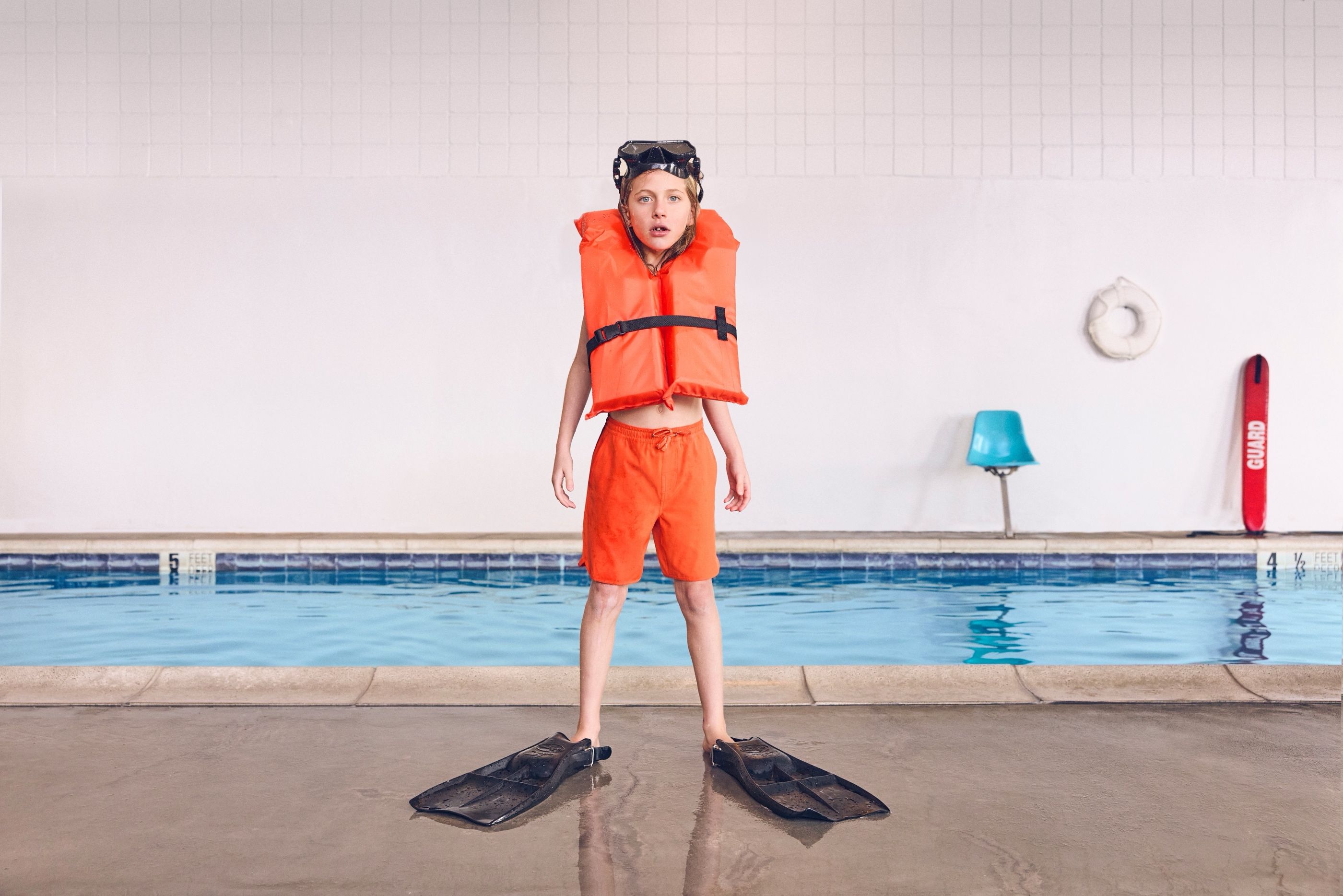 Boy standing at an indoor pool wearing an orange life vest, swim shorts, and fins, with a lifeguard station behind him.