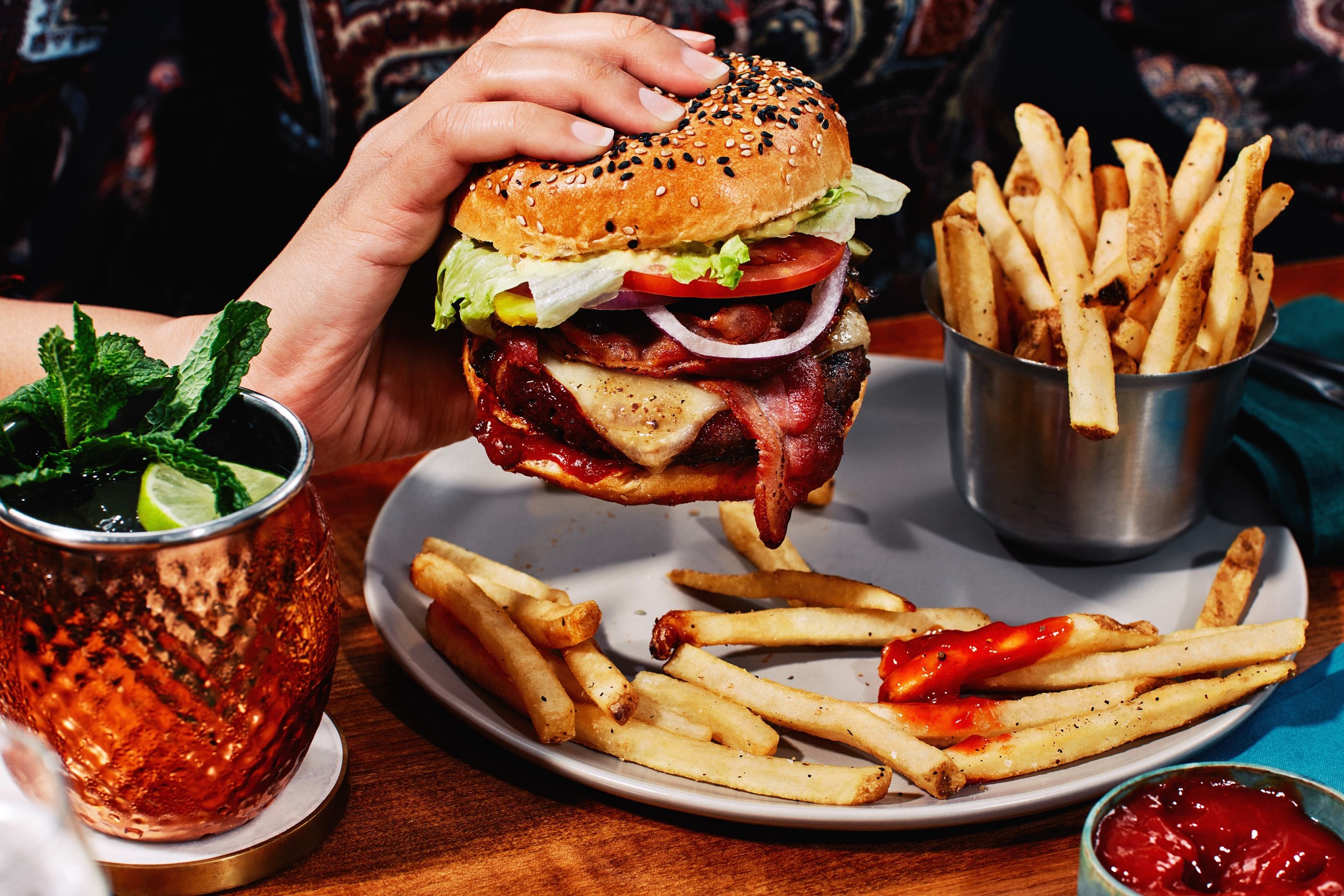 A hand lifts a bacon cheeseburger with lettuce, tomato and onion from a plate of fries, beside a copper mug cocktail.
