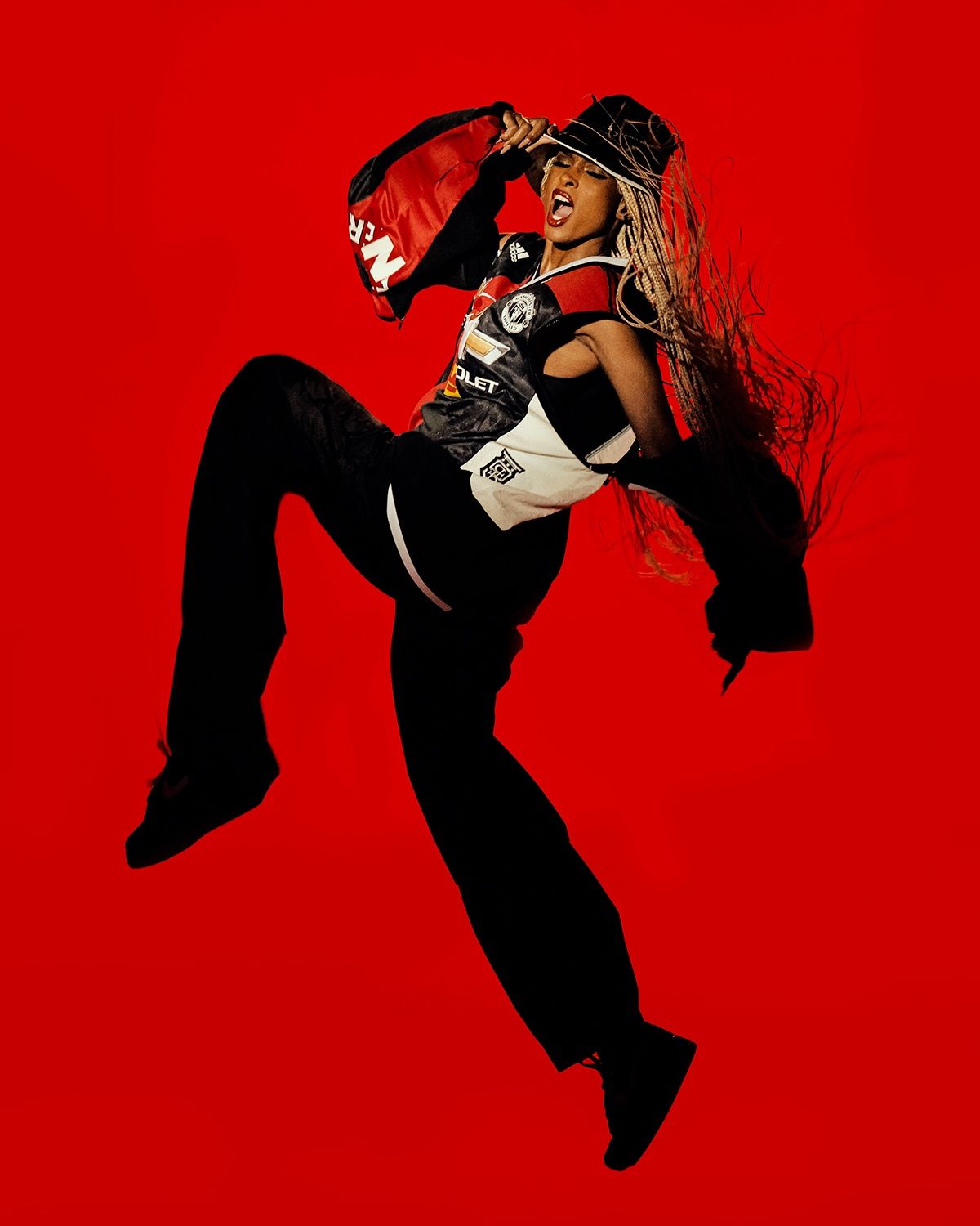 Woman mid-kick wearing a Manchester United jersey and black pants, holding a red jacket, red backdrop.