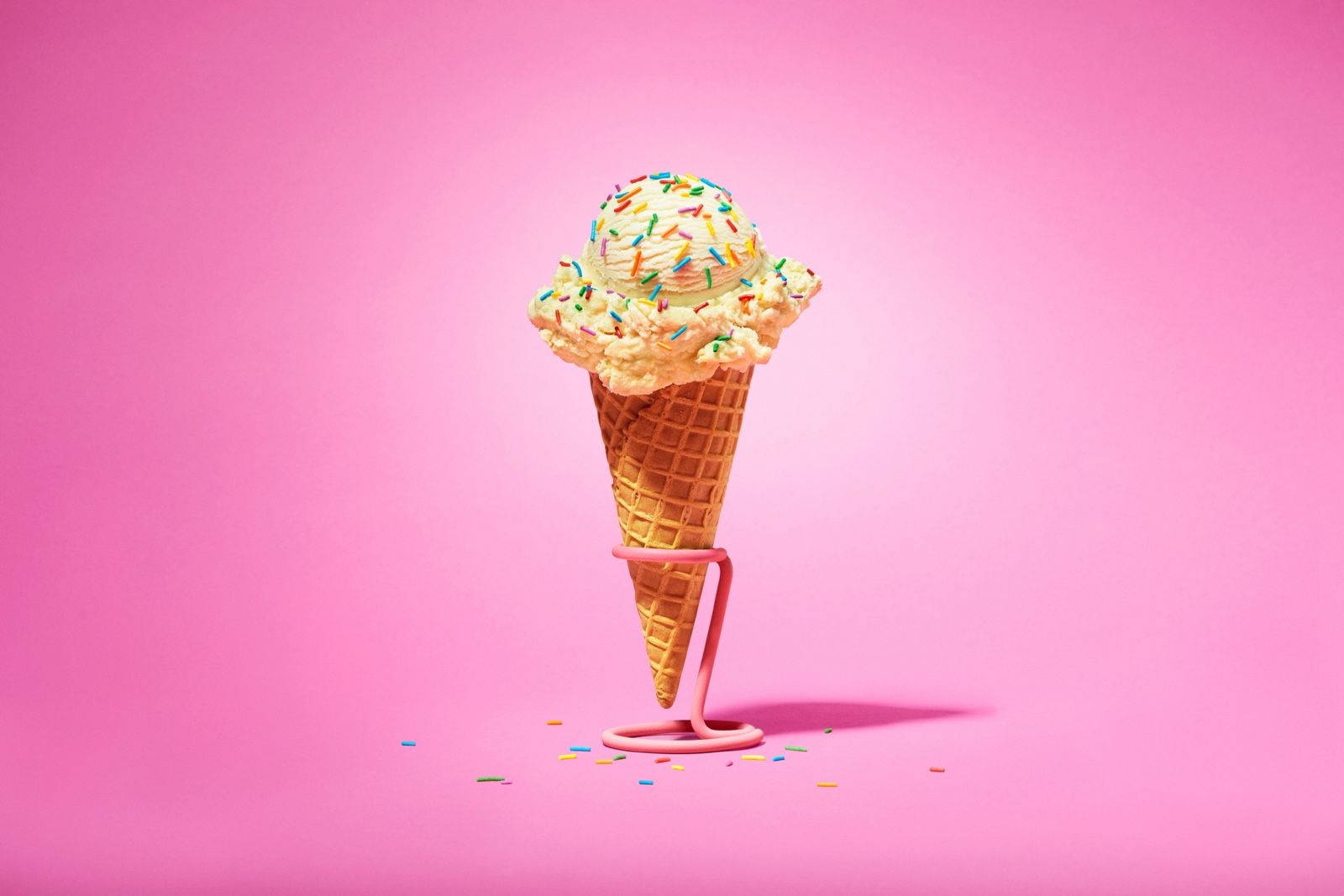 A waffle cone topped with a scoop of vanilla ice cream and rainbow sprinkles, standing in a stand on a pink background.