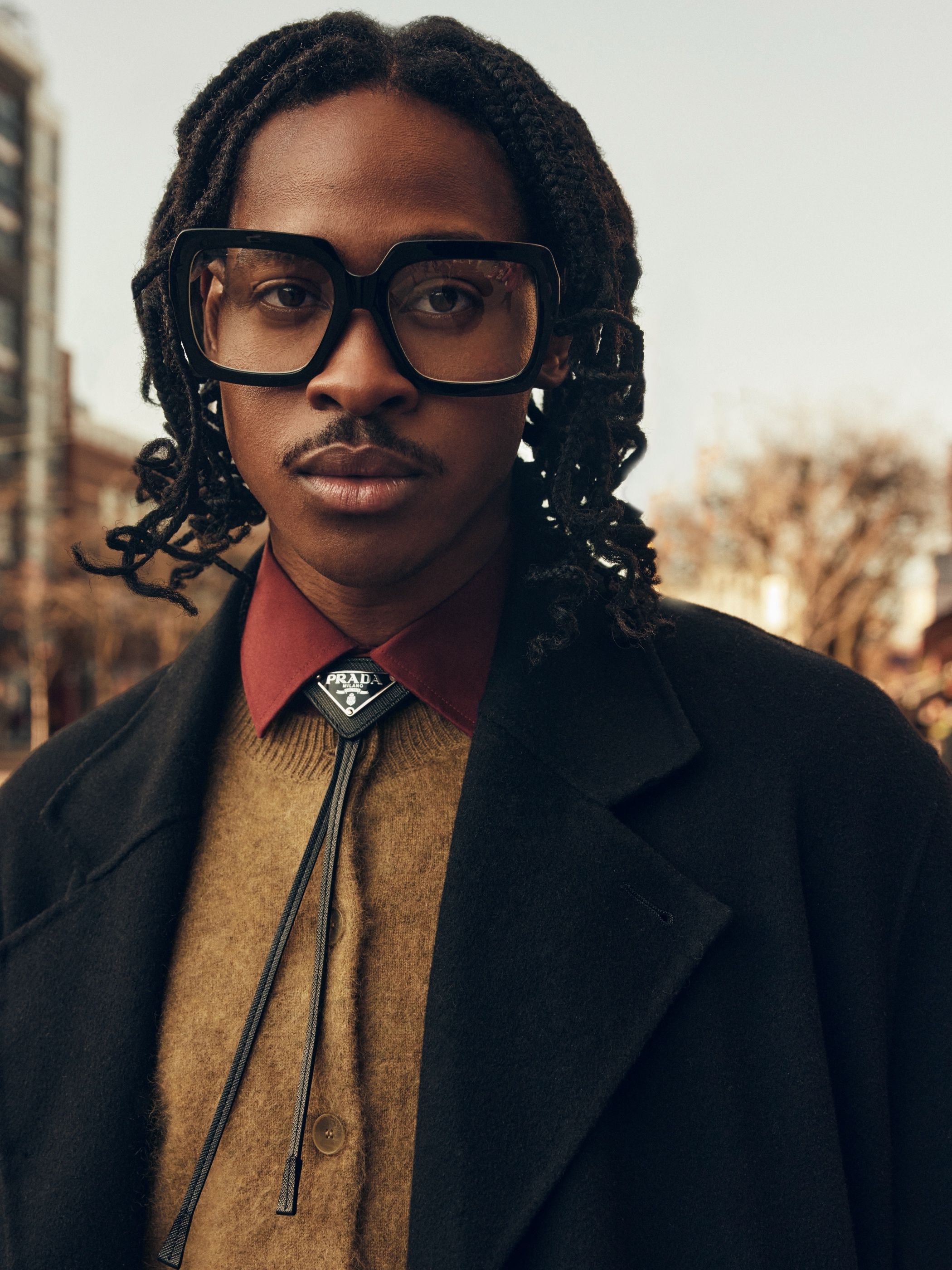 Close-up of a man with braided hair in a black coat and olive sweater, collar fastened with a bolo tie bearing a Prada tag.