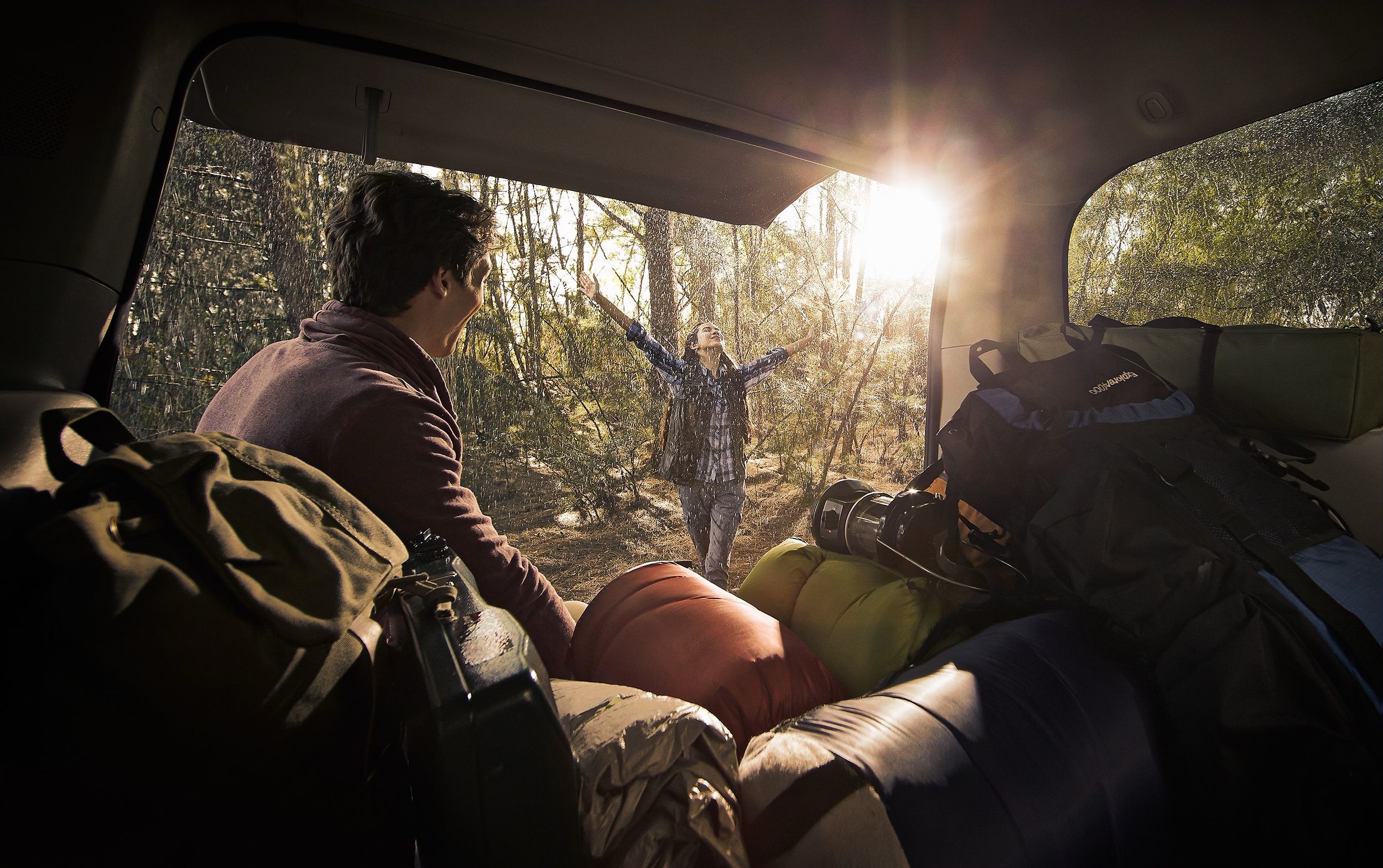View from inside a vehicle's trunk of a man watching a woman raise her arms in the rain in a forest clearing.