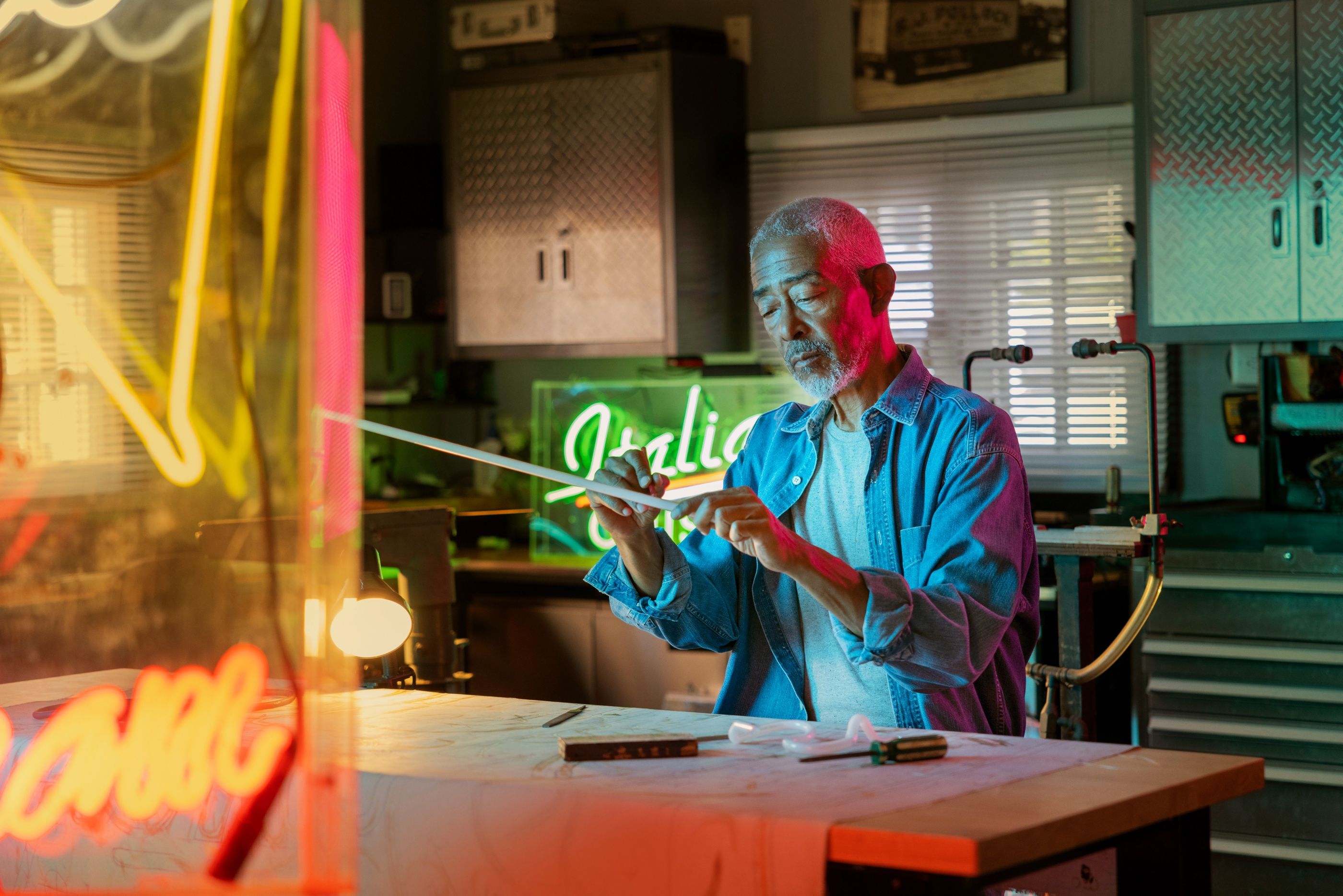 Older man in a workshop shapes a glowing neon tube surrounded by colorful signage.