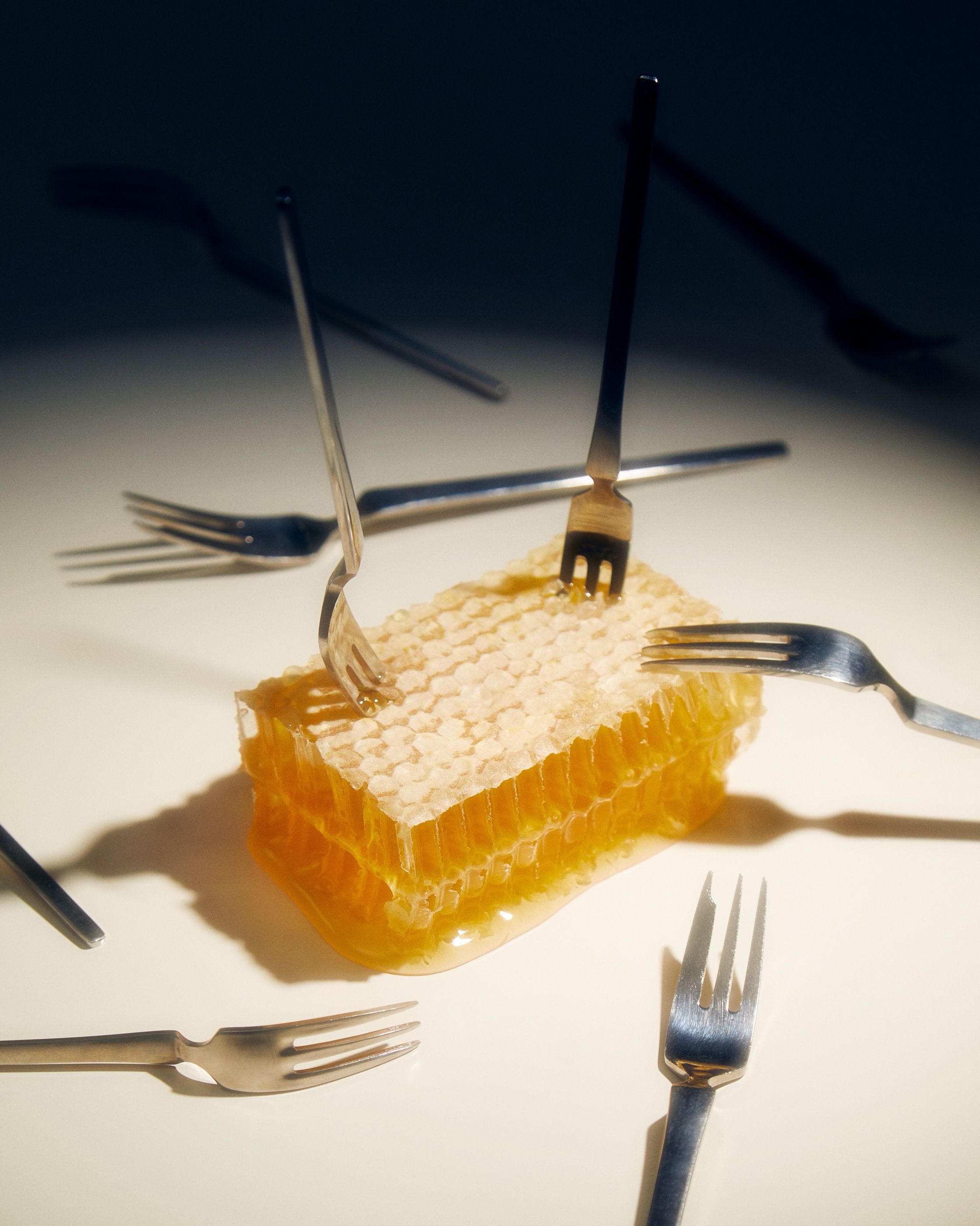 A block of honeycomb dripping honey, surrounded by forks stuck in at odd angles on a white surface.