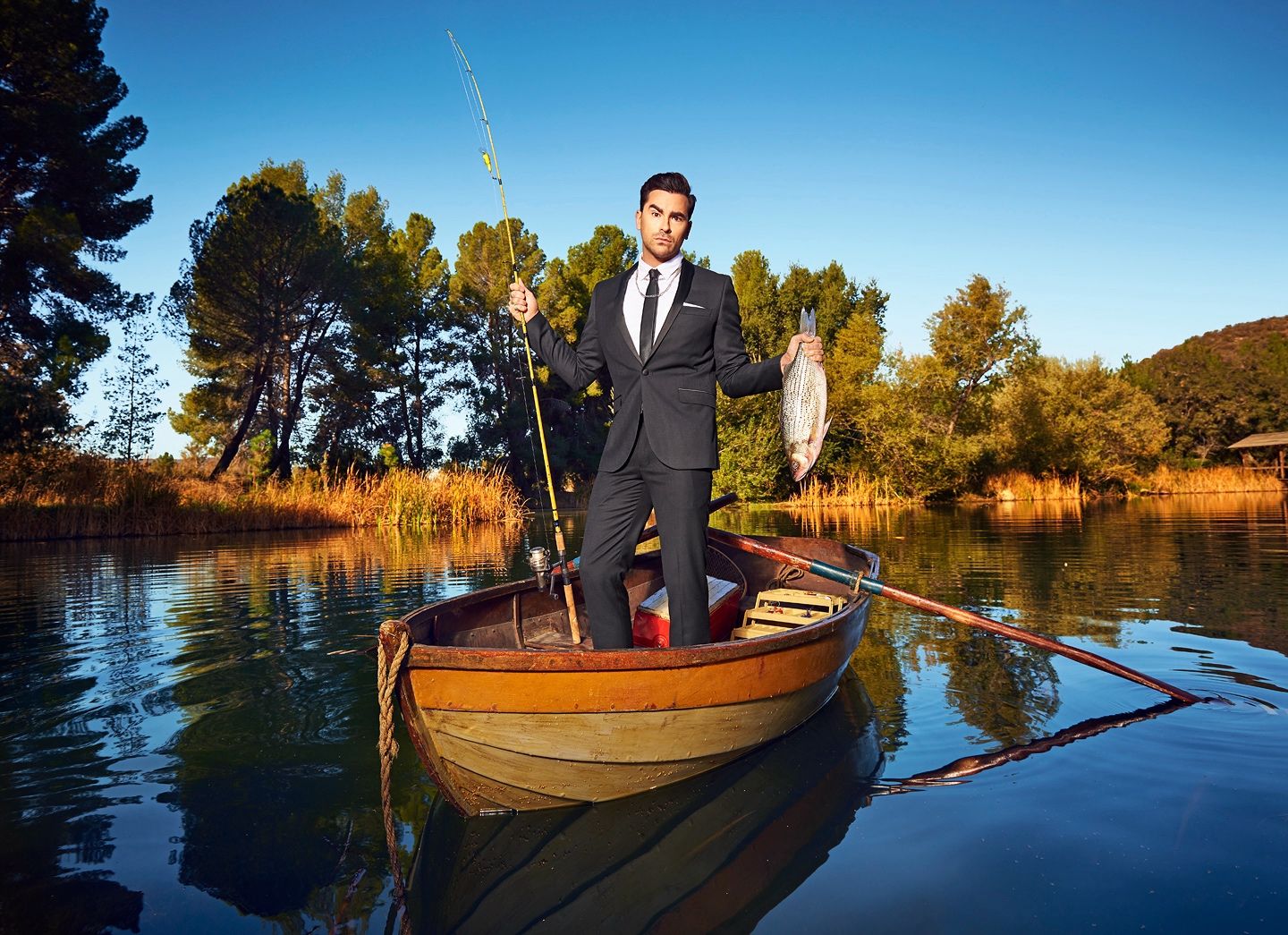 Man in a suit stands in a wooden rowboat on a lake, holding a fishing rod and a caught fish.