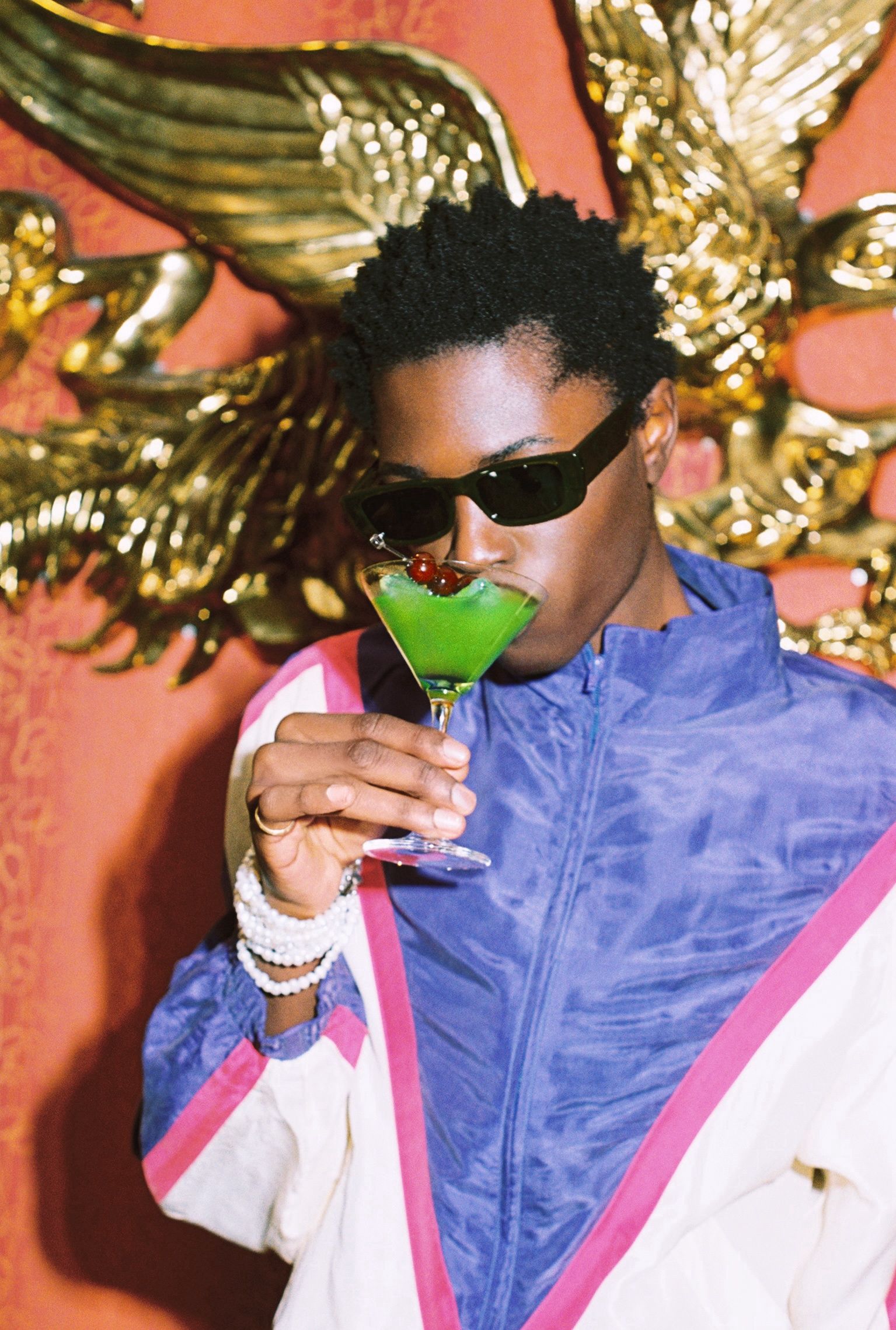 Man in sunglasses and a colorblock jacket drinking a green cocktail garnished with cherries.