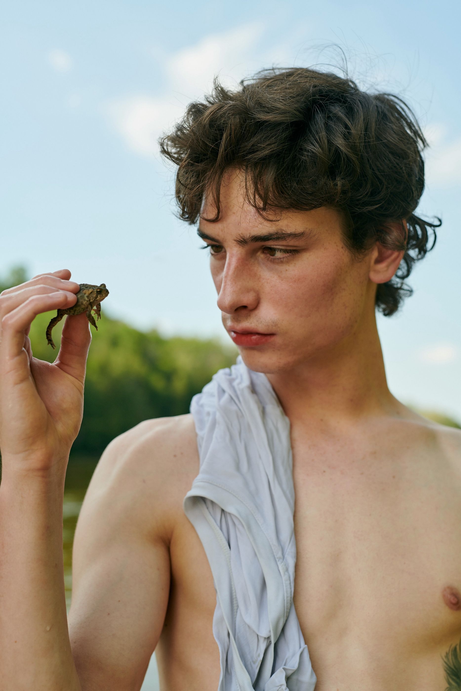 Young man outdoors holding a small toad up near his face, shirtless with a light cloth draped over one shoulder.