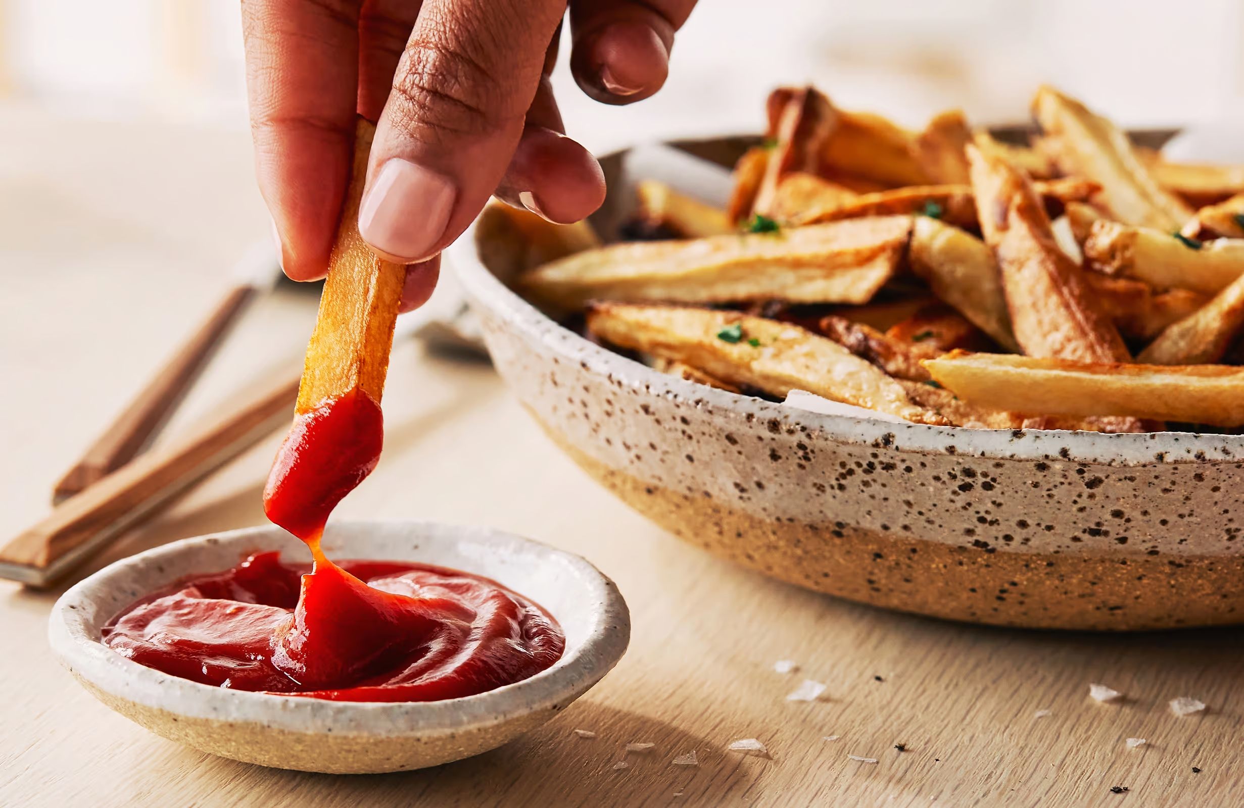 A hand dips a french fry into a dish of ketchup beside a bowl of seasoned fries on a wood table.