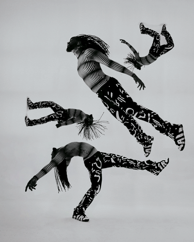 Three dancers in patterned black-and-white bodysuits captured mid-air in acrobatic poses, grey backdrop.