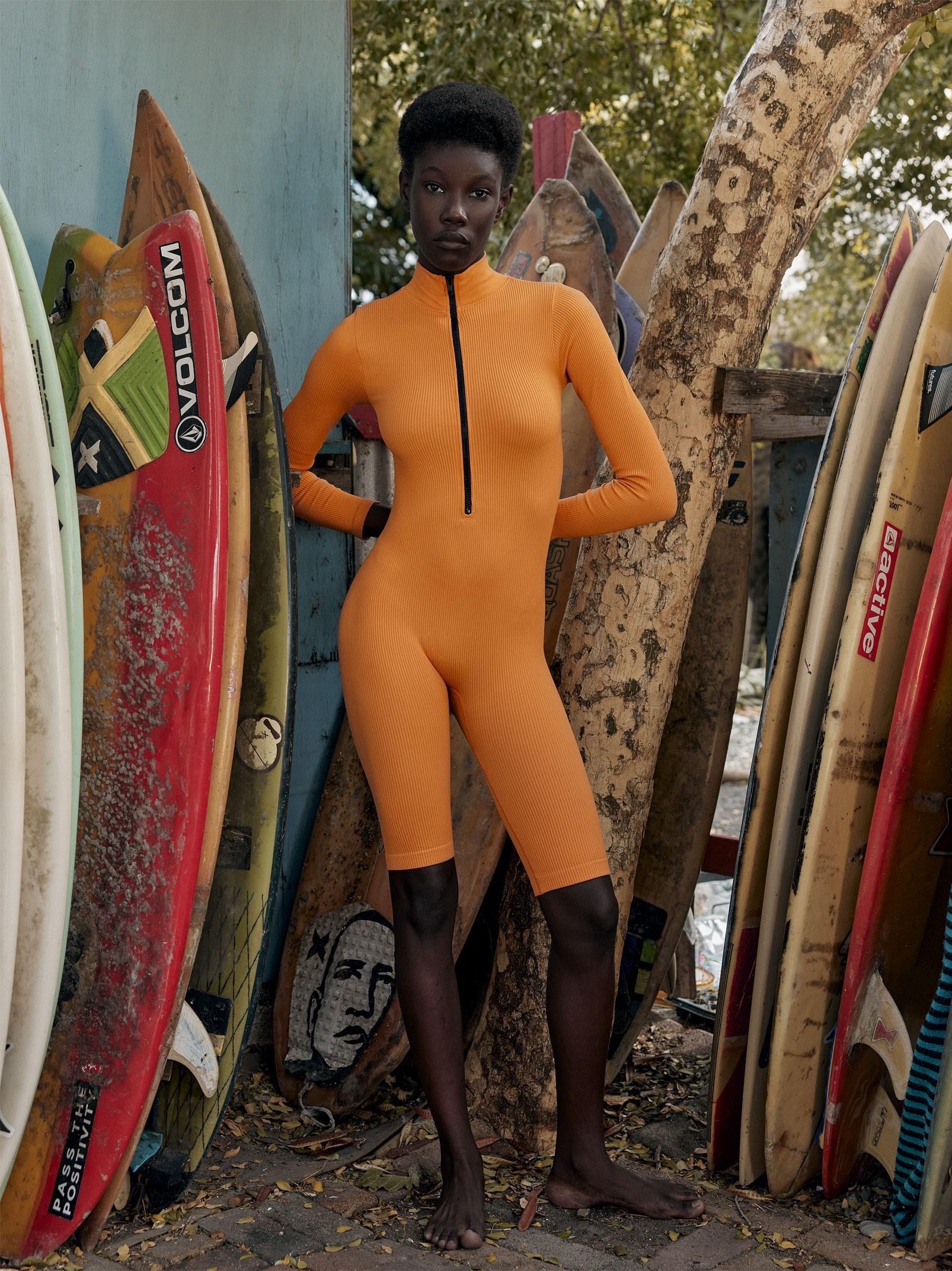 Woman in an orange ribbed zip-front catsuit stands barefoot among surfboards leaning against a wall and tree.