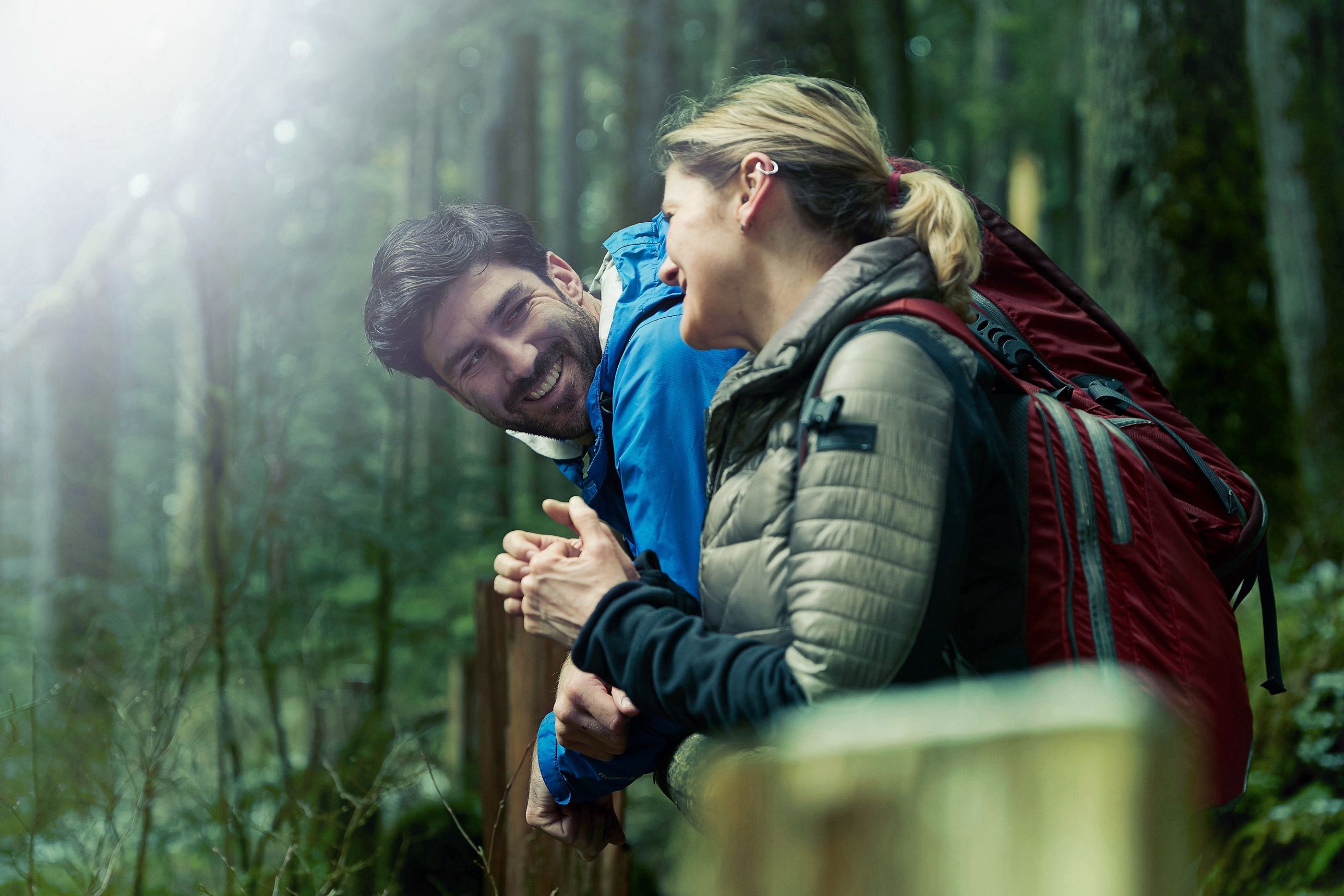Man and woman lean on a wooden rail in a forest, laughing together, the woman wearing a large backpack.