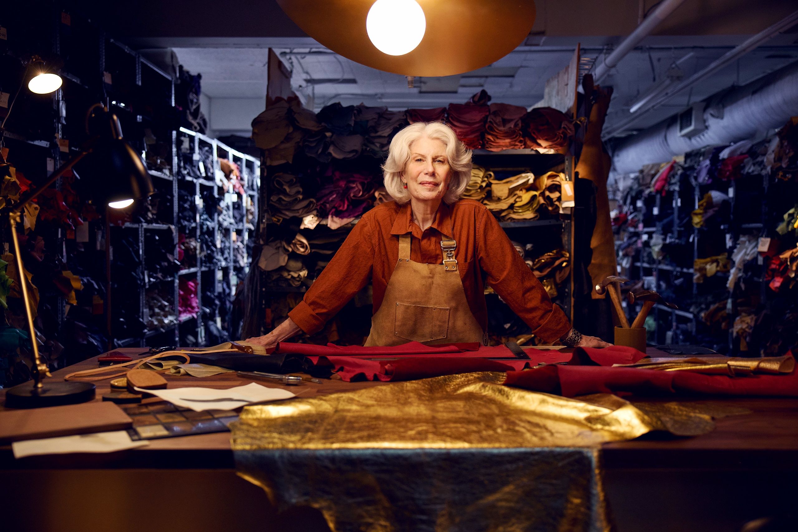 Woman in an apron stands at a leather workshop table surrounded by shelves of hides.