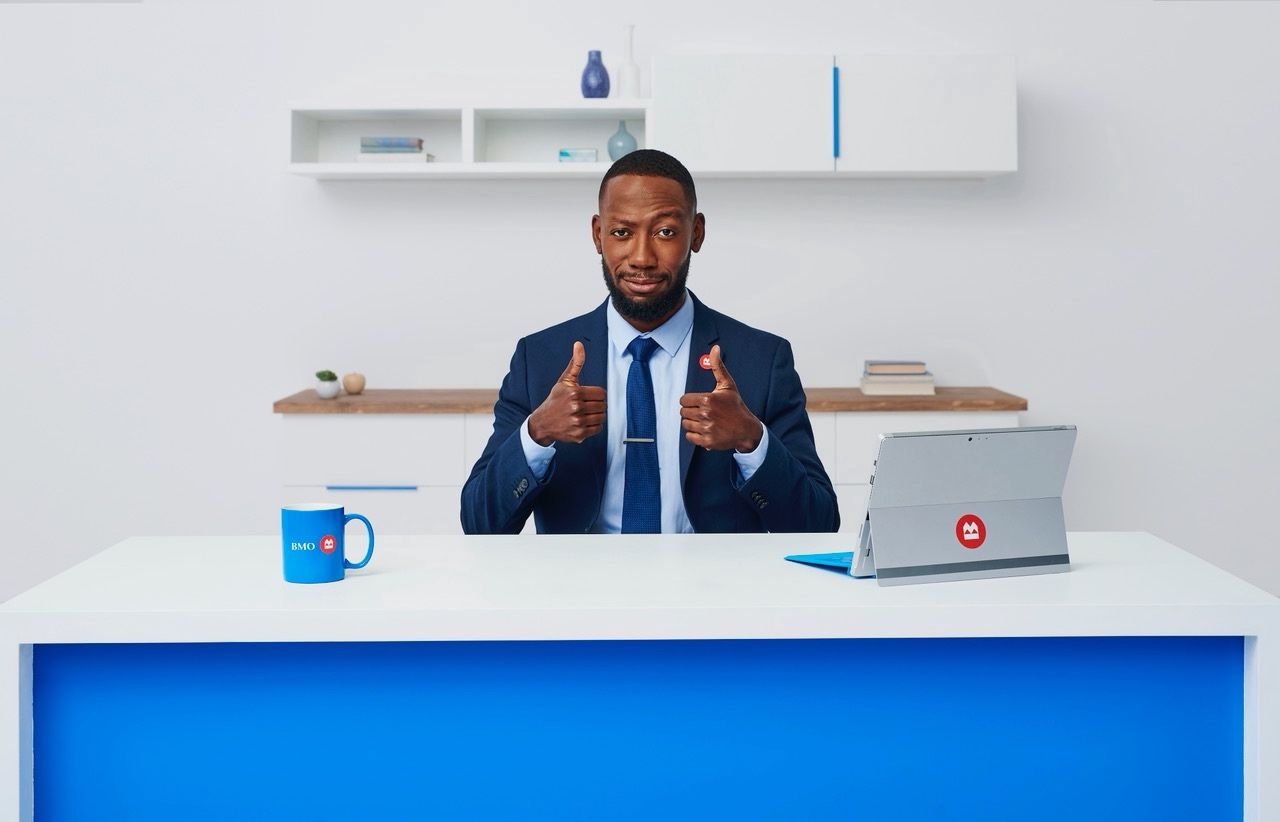 Man in a navy suit gives two thumbs up at a desk with a BMO mug and a tablet bearing the BMO logo.