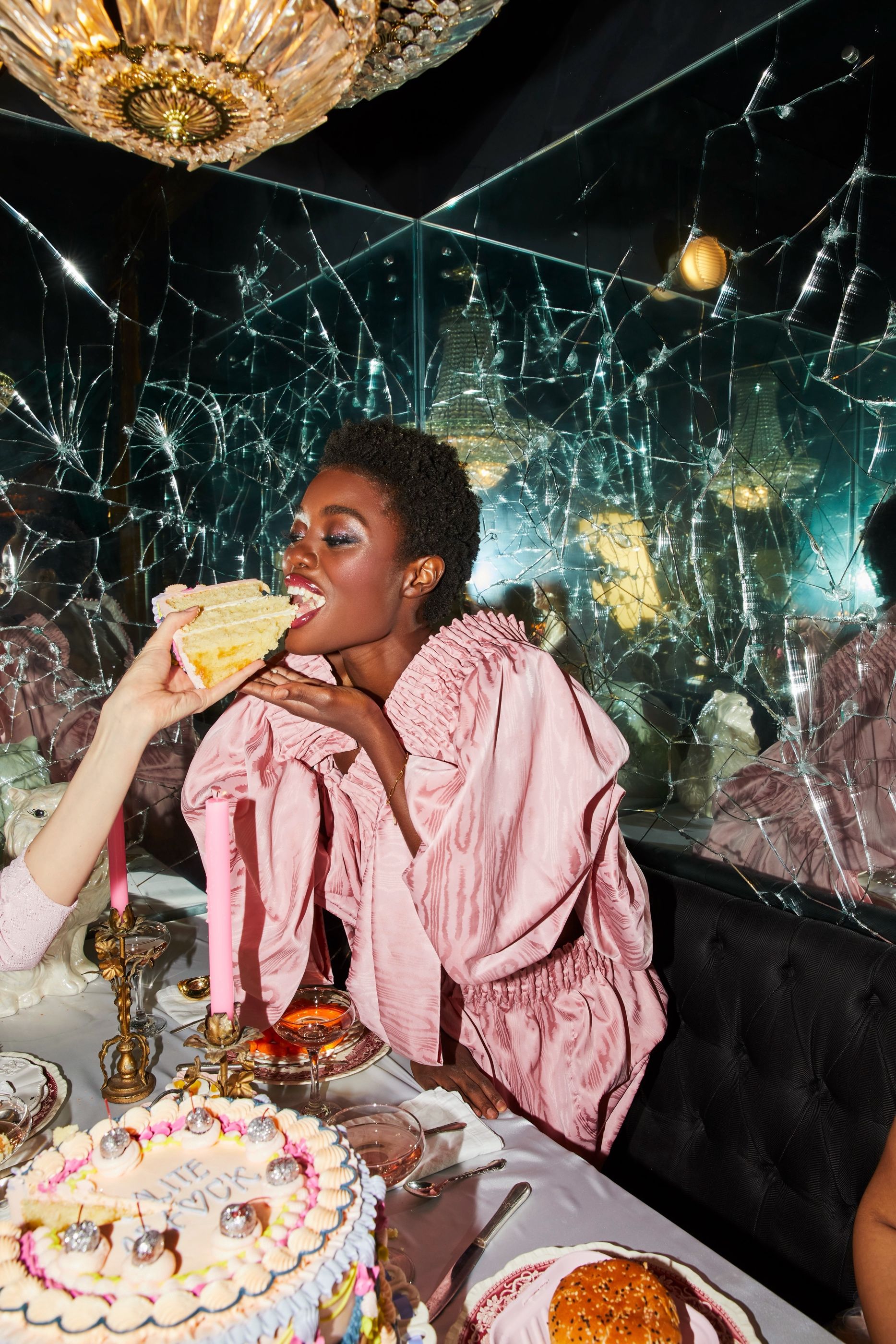 Woman in a pink ruffled outfit biting into a slice of birthday cake at a table, shattered-mirror wall behind her.