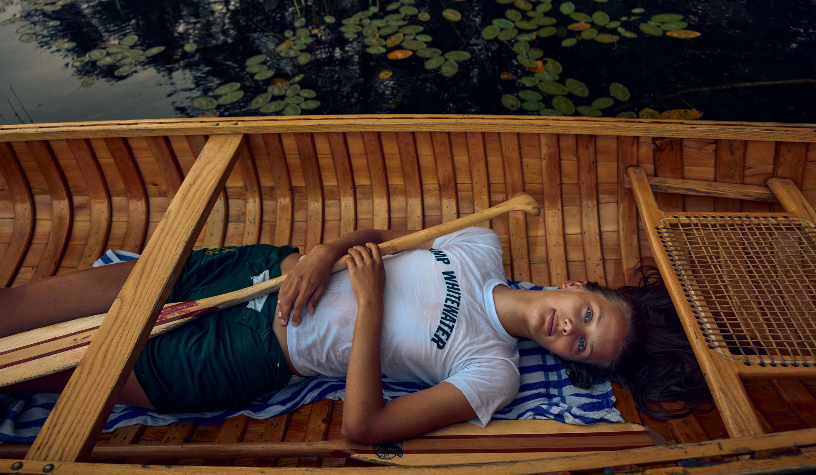 Girl lying in a wooden canoe holding an oar, wearing a Camp Whitewater T-shirt, near lily pads on the water.
