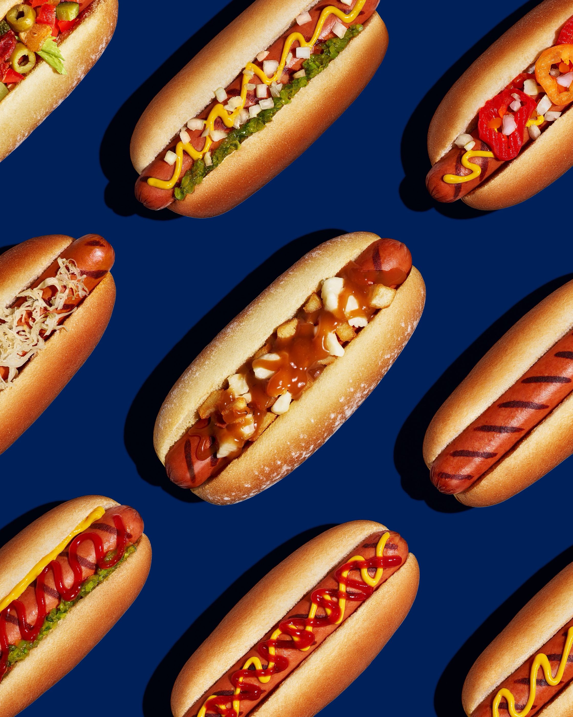 Assorted hot dogs with toppings including mustard, onion, sauerkraut and peppers, arranged on a blue surface.