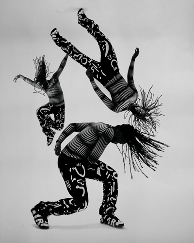 Three dancers in patterned black-and-white bodysuits mid-flip, one kneeling, grey backdrop.