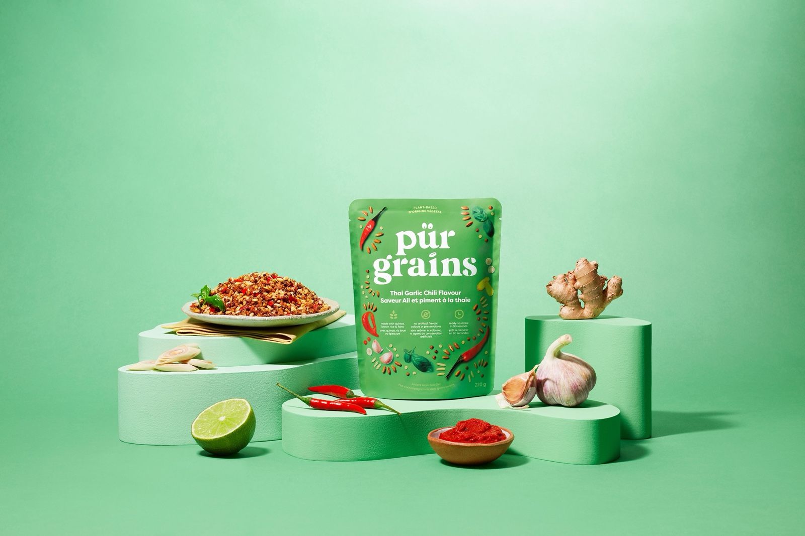 Pür Grains Thai Garlic Chili grain pouch on green podiums with a bowl of grain salad, chilies, garlic, ginger and lime.