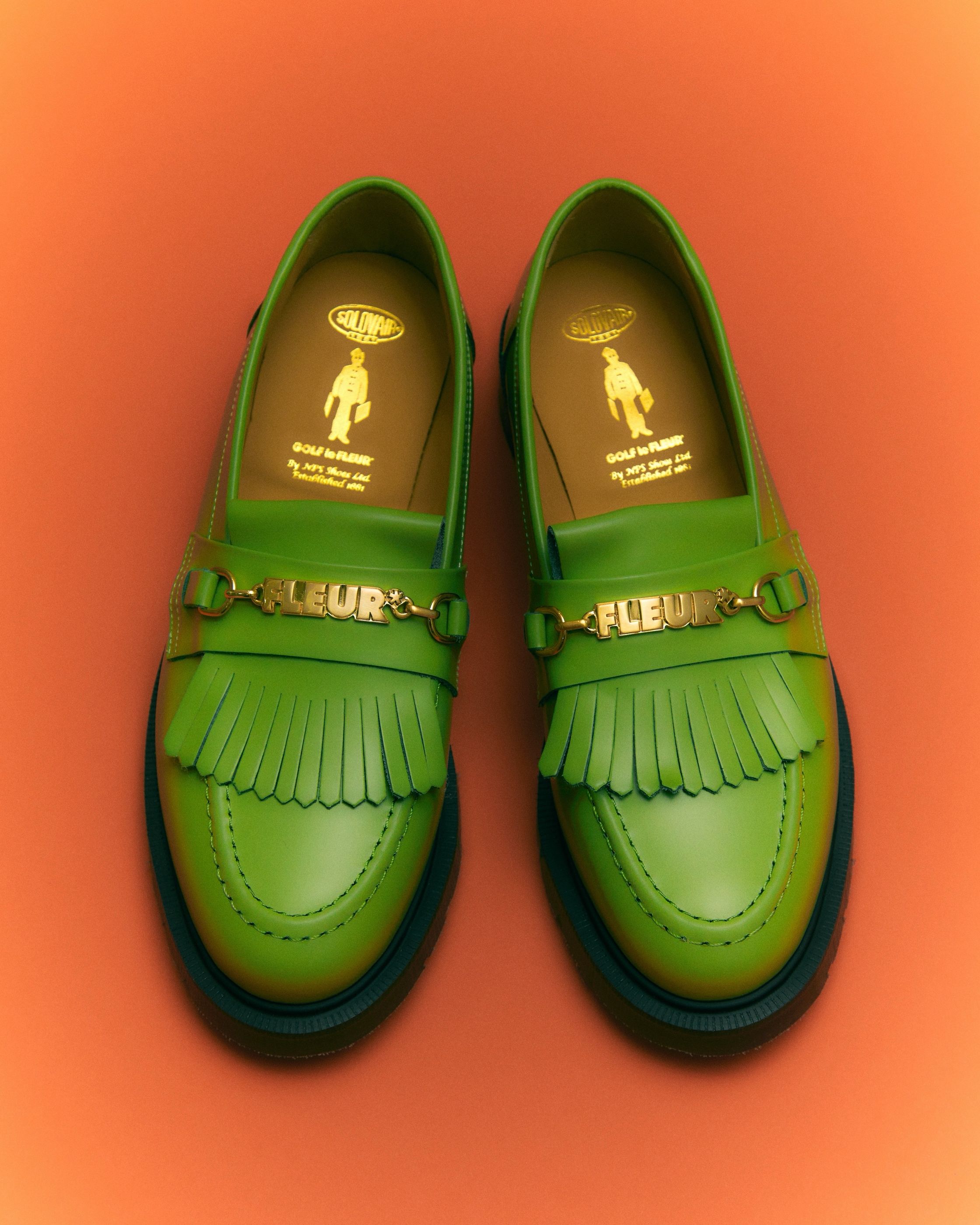 Green leather tasseled loafers with gold FLEUR hardware, by Solovair, on an orange background.