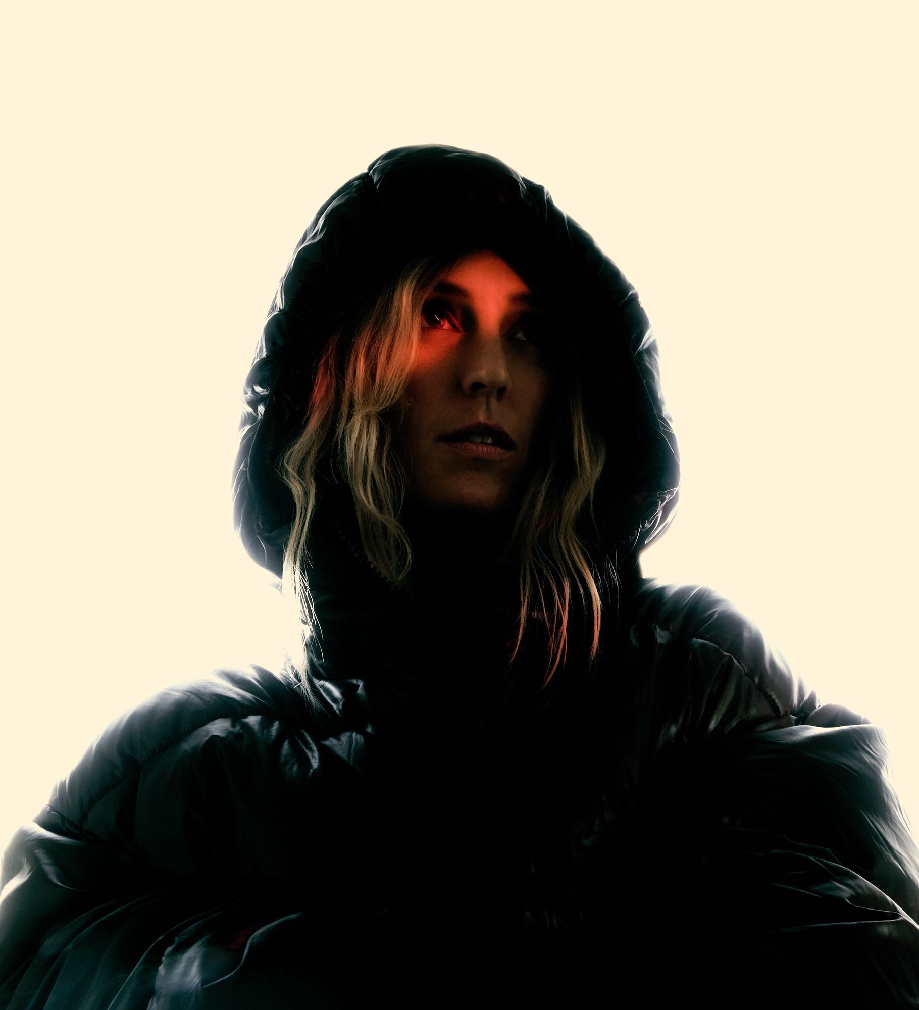 Person in a dark hooded jacket with red light across the eyes, cream background.