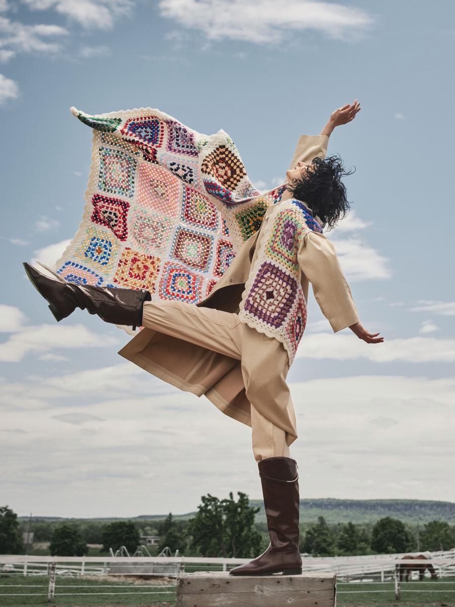 Person in a tan suit with a colorful crochet vest kicks one leg back while balancing on a wooden block in a grassy paddock.