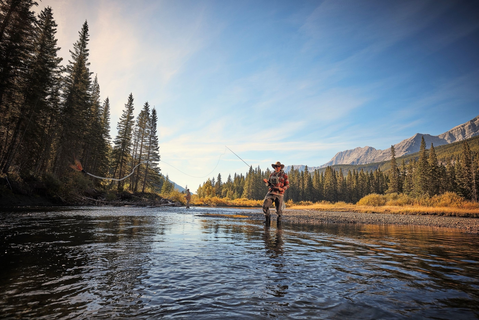 Man fly-fishing in a river casts his line, mountains and pine trees behind him and another angler downstream.
