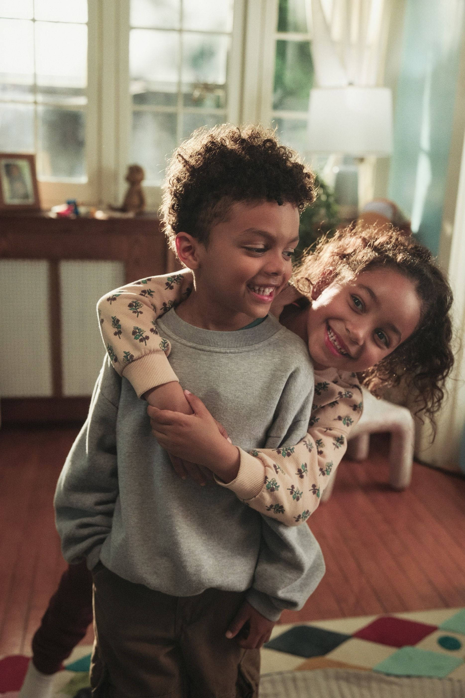 Two children hug and smile together in a sunlit living room.