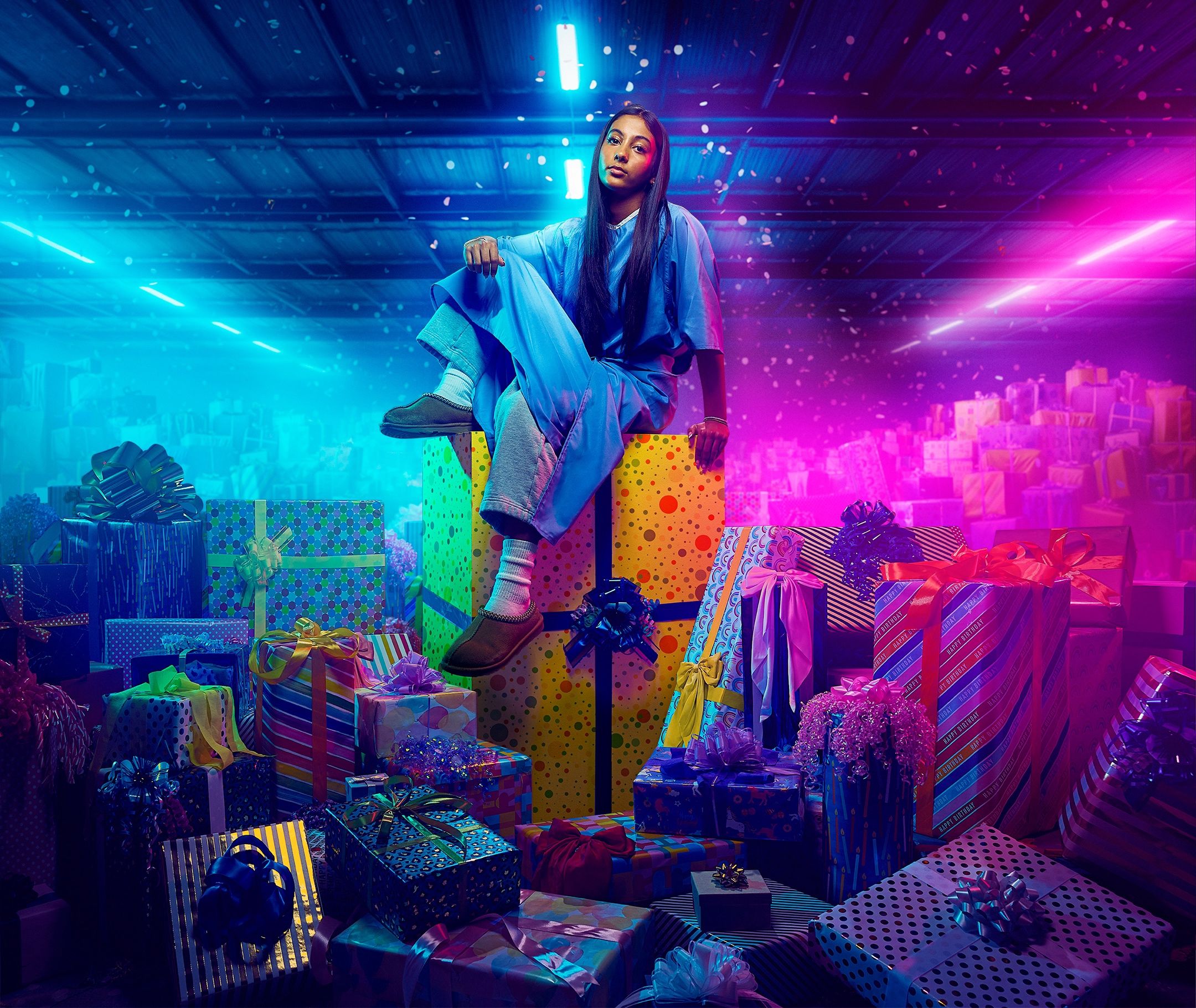Girl in blue loungewear sits atop wrapped birthday gifts under blue and pink neon warehouse lighting.