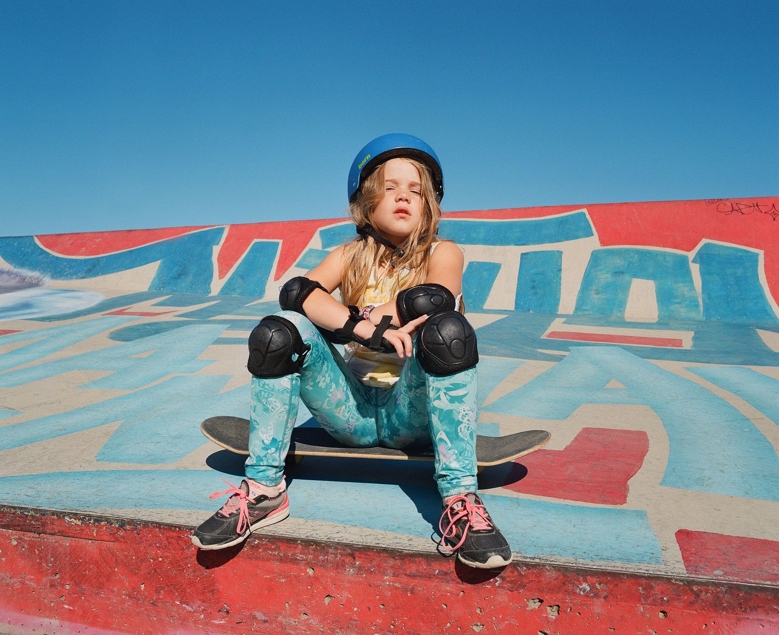 Girl sitting on a skateboard wearing a helmet and knee pads in front of a graffiti-covered ramp, eyes closed in sunlight.