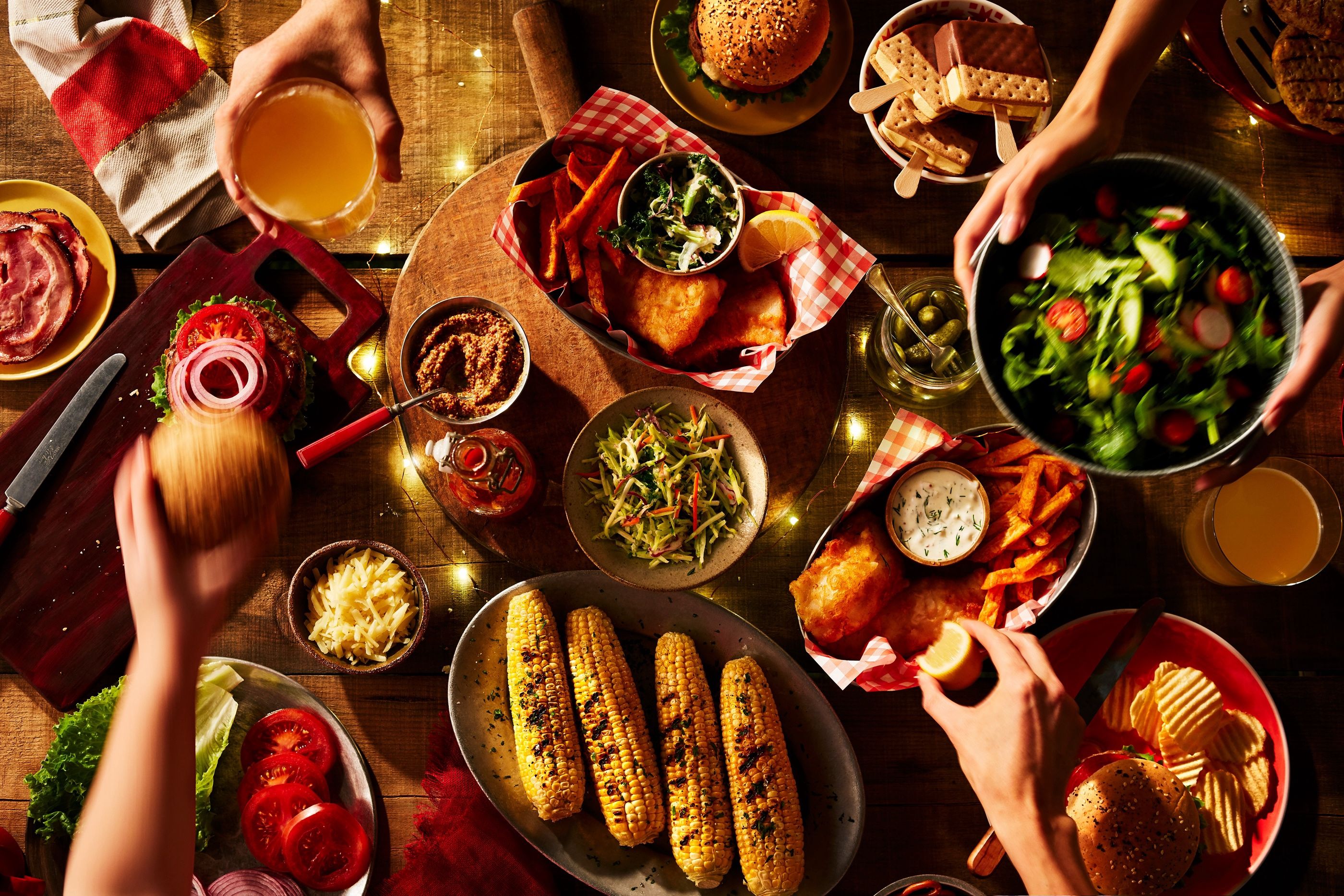 Overhead view of a spread with burgers, fish and chips, grilled corn and salad, with hands reaching in.