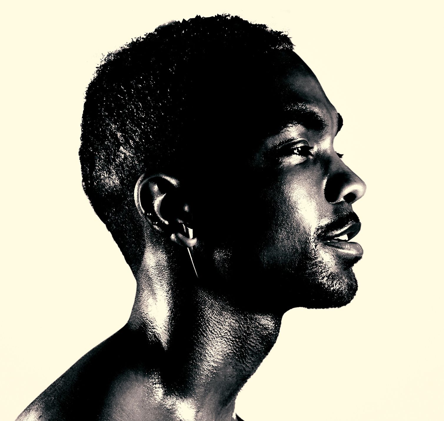 Profile portrait of a man with short hair and a spike earring, bare shoulders, cream background.