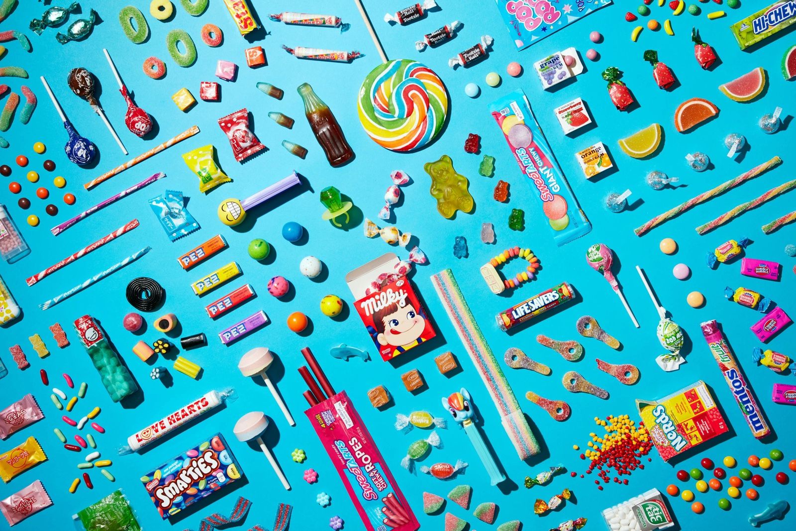 An assortment of candies including Smarties, Nerds, Mentos, PEZ, Life Savers and gummy bears arranged on a blue background.