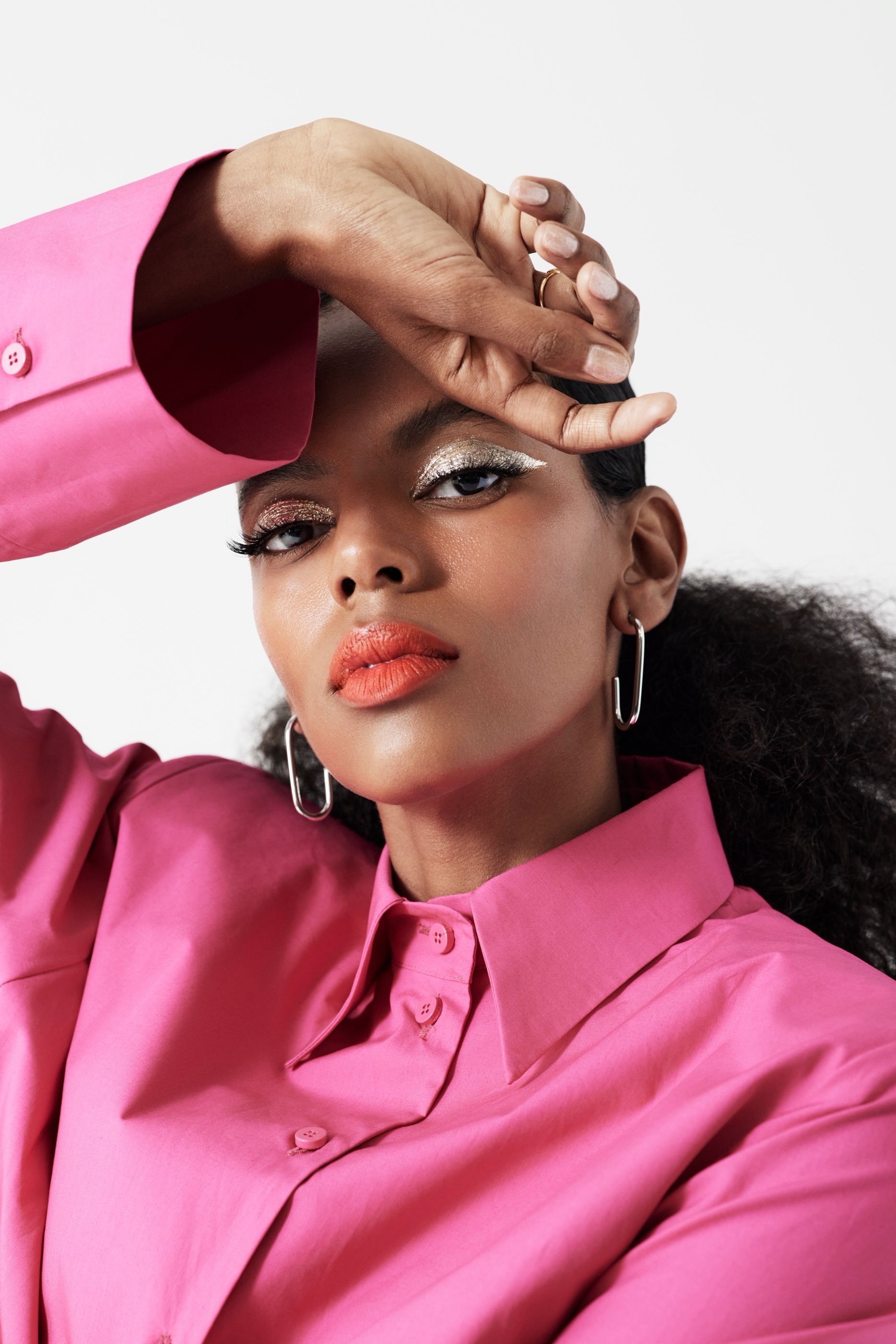 Close-up of a woman in a pink shirt with glitter eyeshadow and orange lipstick, one hand raised to her forehead.