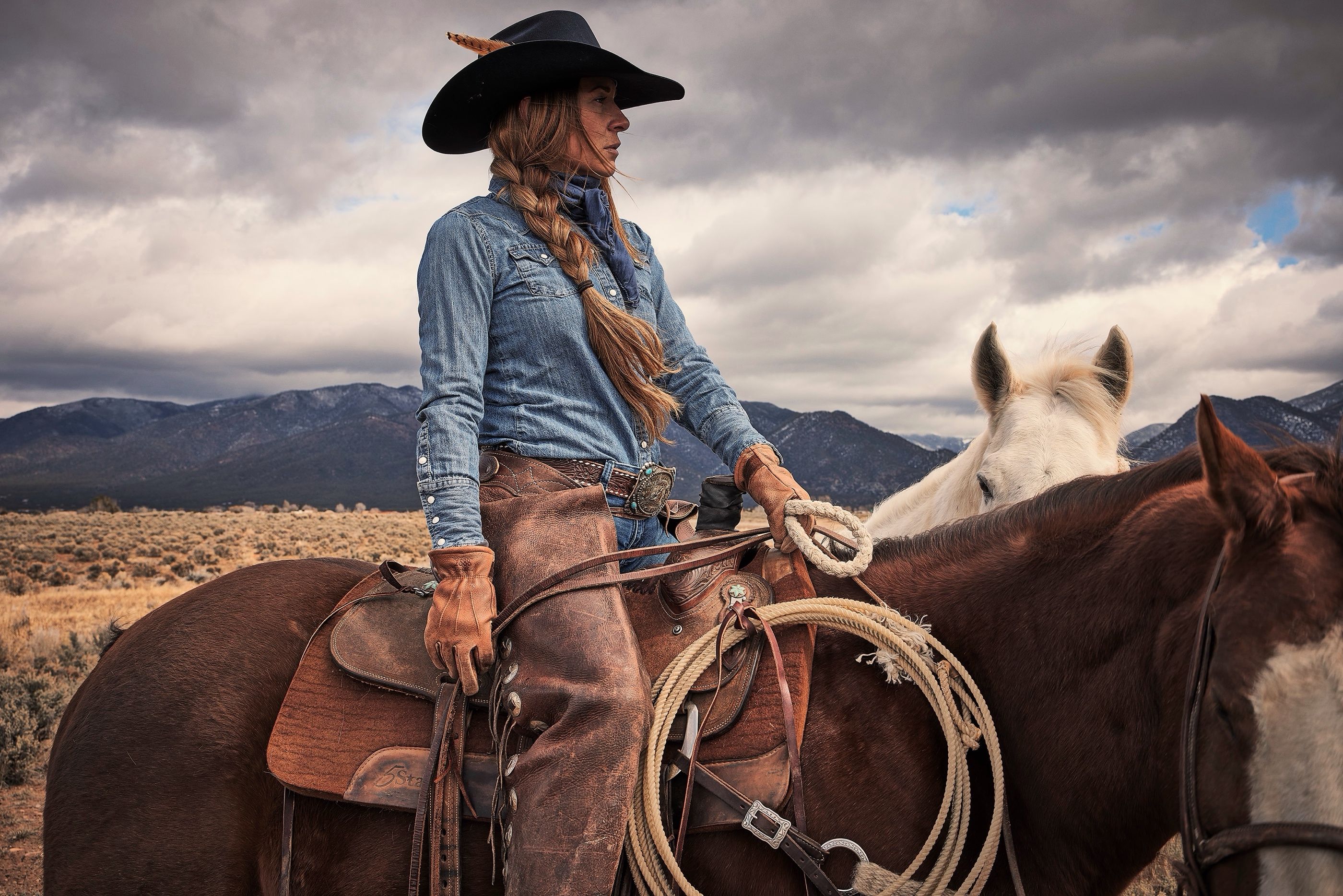 Woman in a black cowboy hat and denim shirt sits on a horse holding a coiled rope, mountains and clouds behind her.