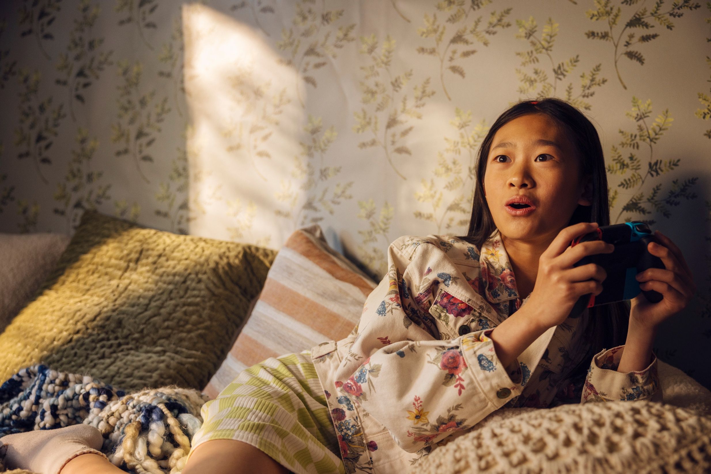 A young girl sits comfortably on a sofa in a warmly lit room, holding a video game controller. She wears a floral button-up shirt and green-and-white striped shorts, surrounded by textured pillows and blankets. Floral wallpaper and soft sunlight create a cozy atmosphere