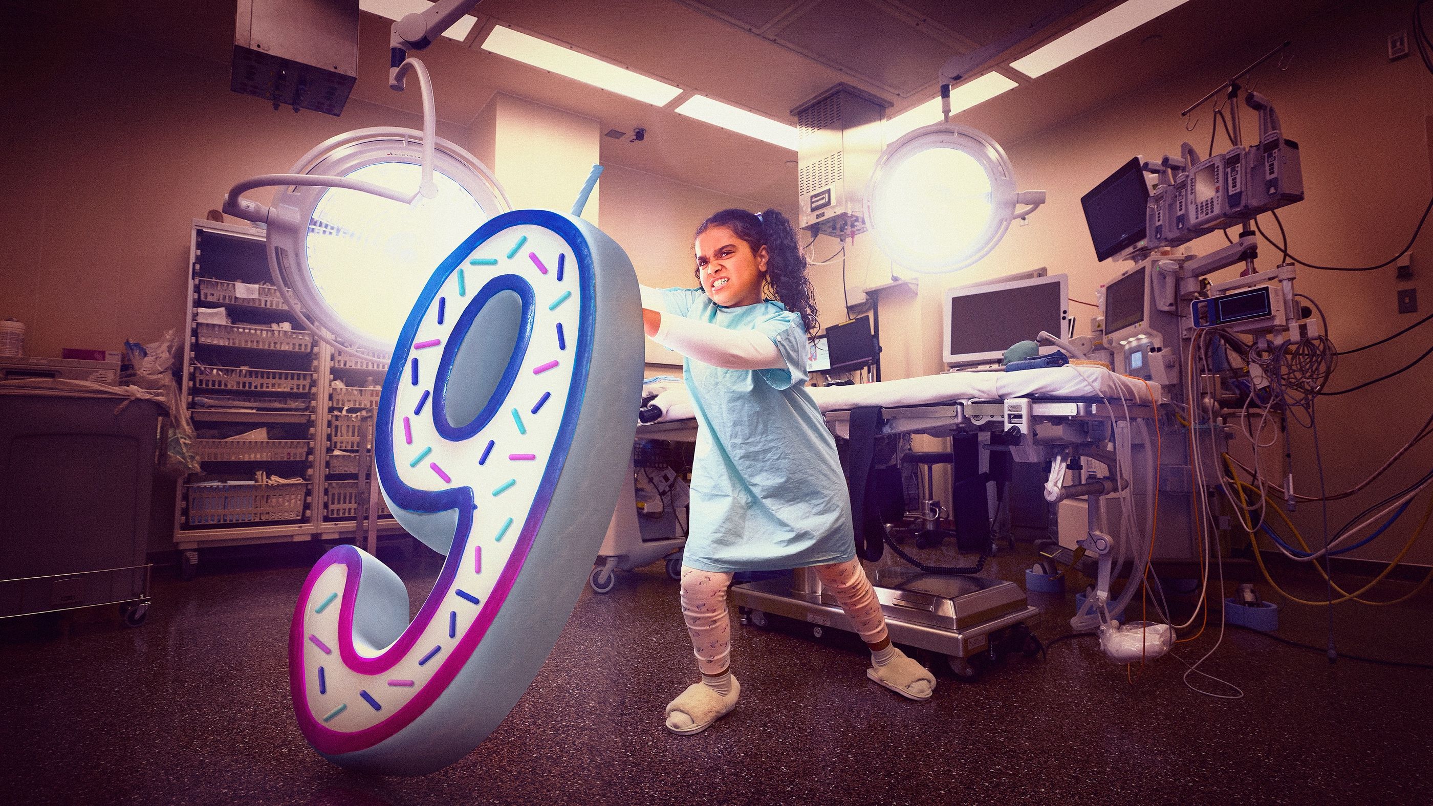 Girl in a hospital gown play-punches a large foam number nine sculpture in a hospital procedure room.