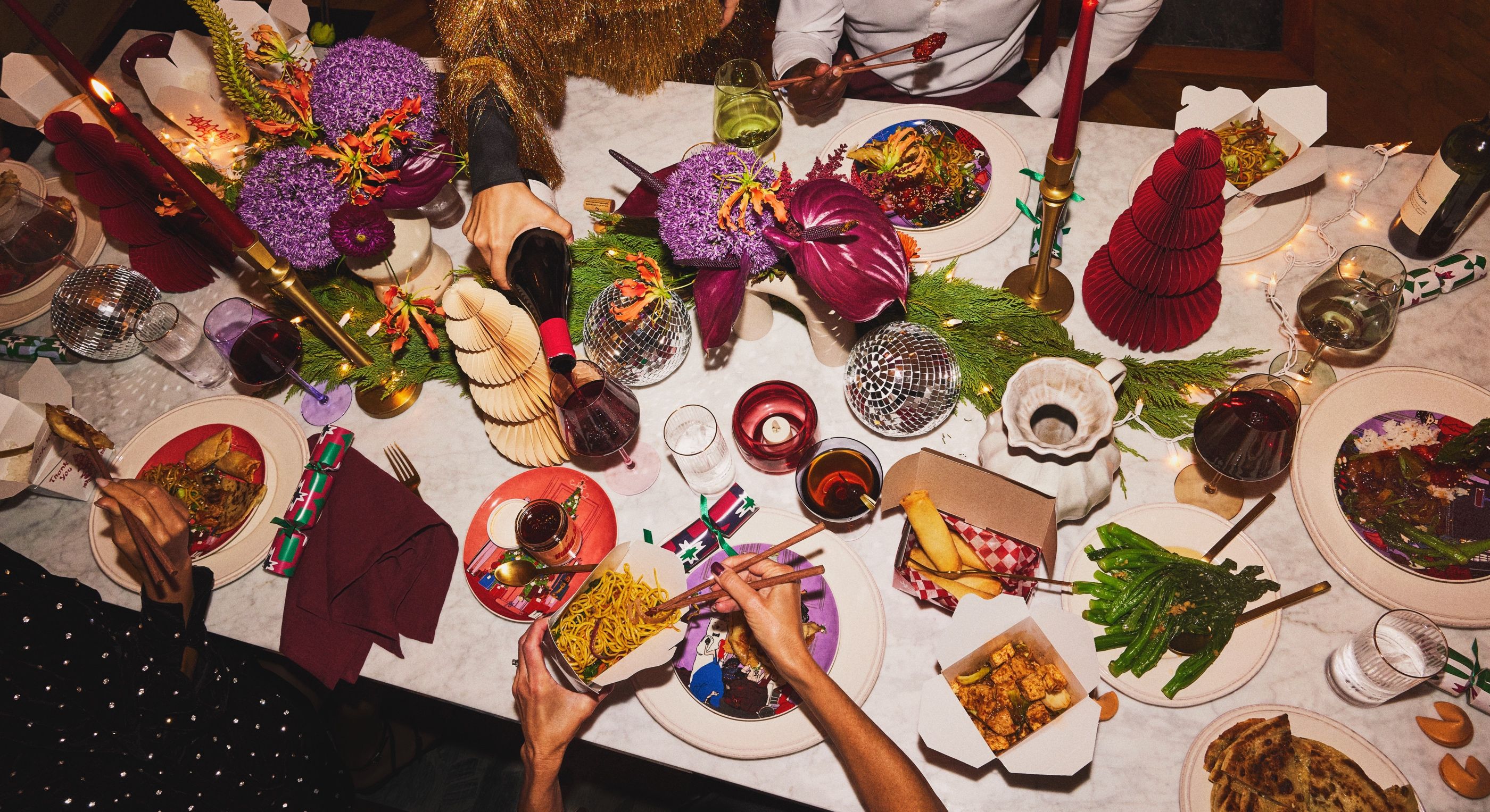 Overhead view of a festive holiday dinner table with takeout containers, noodles, candles, and a floral centerpiece.
