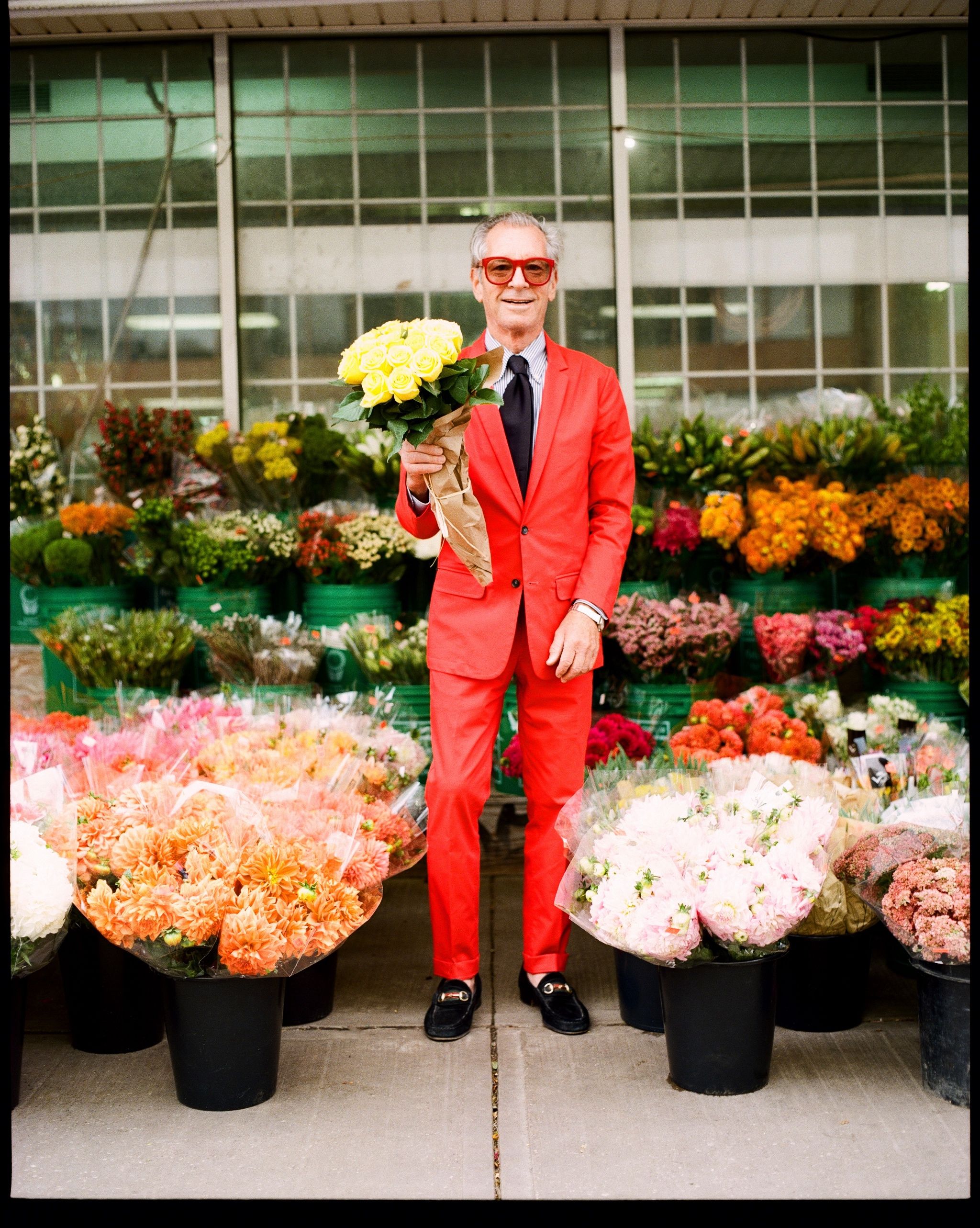 Older man in a red suit and red sunglasses holding a bouquet of yellow roses at an outdoor flower market.