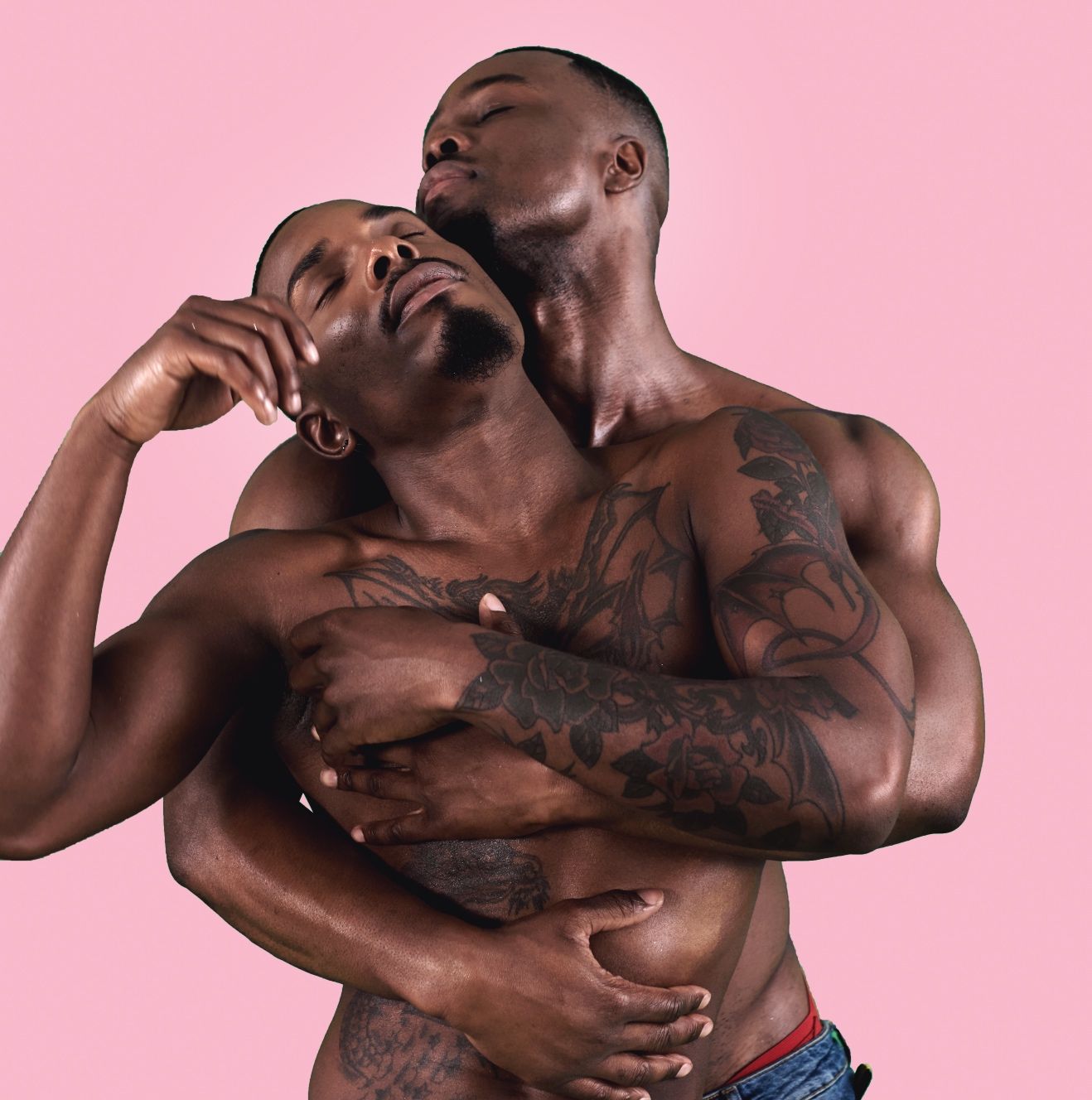 Two shirtless men embracing, one with floral tattoos on his arm and chest, pink background.