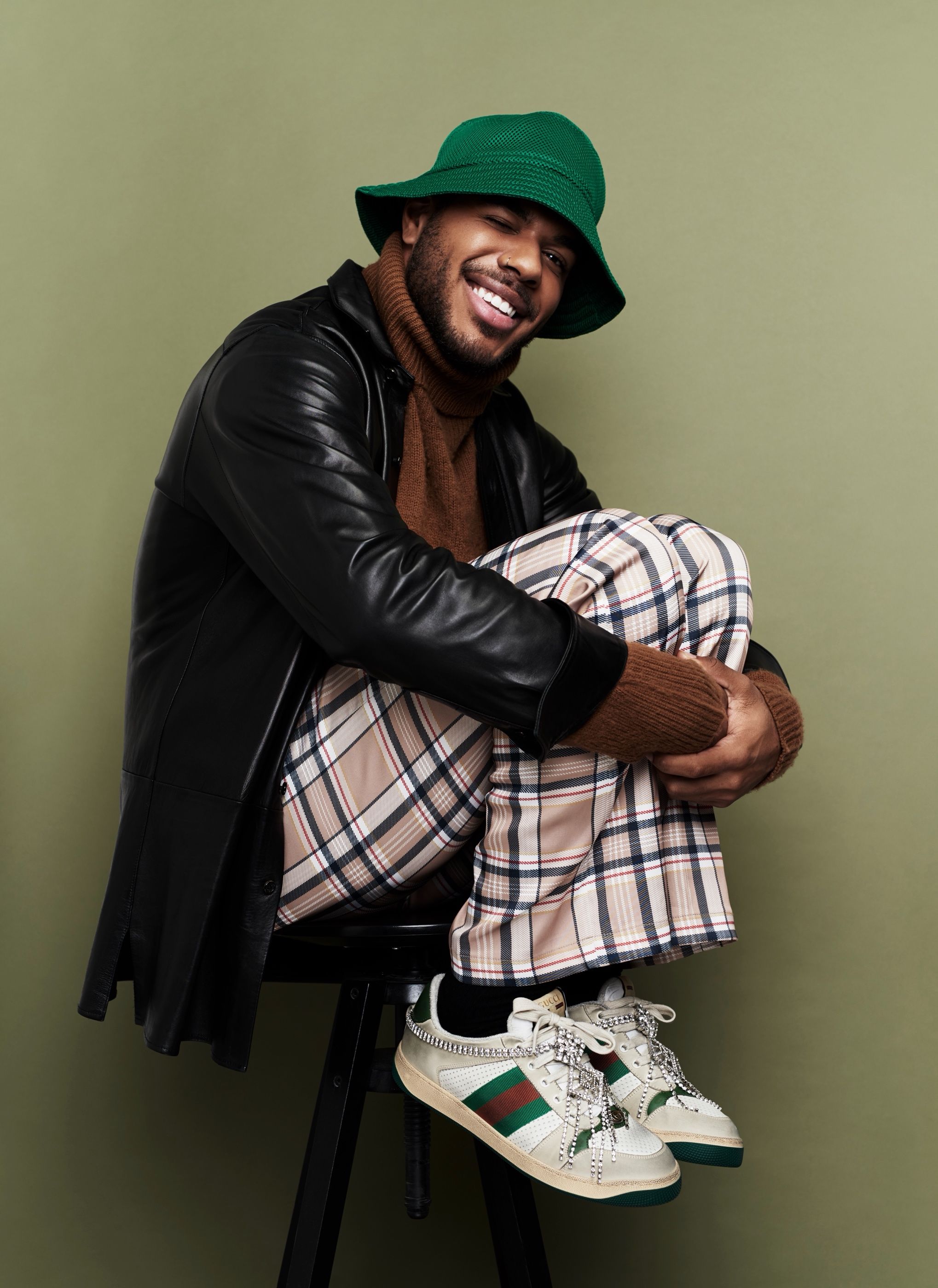 Man in a green bucket hat and black leather jacket sits on a stool wearing plaid trousers and crystal Gucci sneakers.